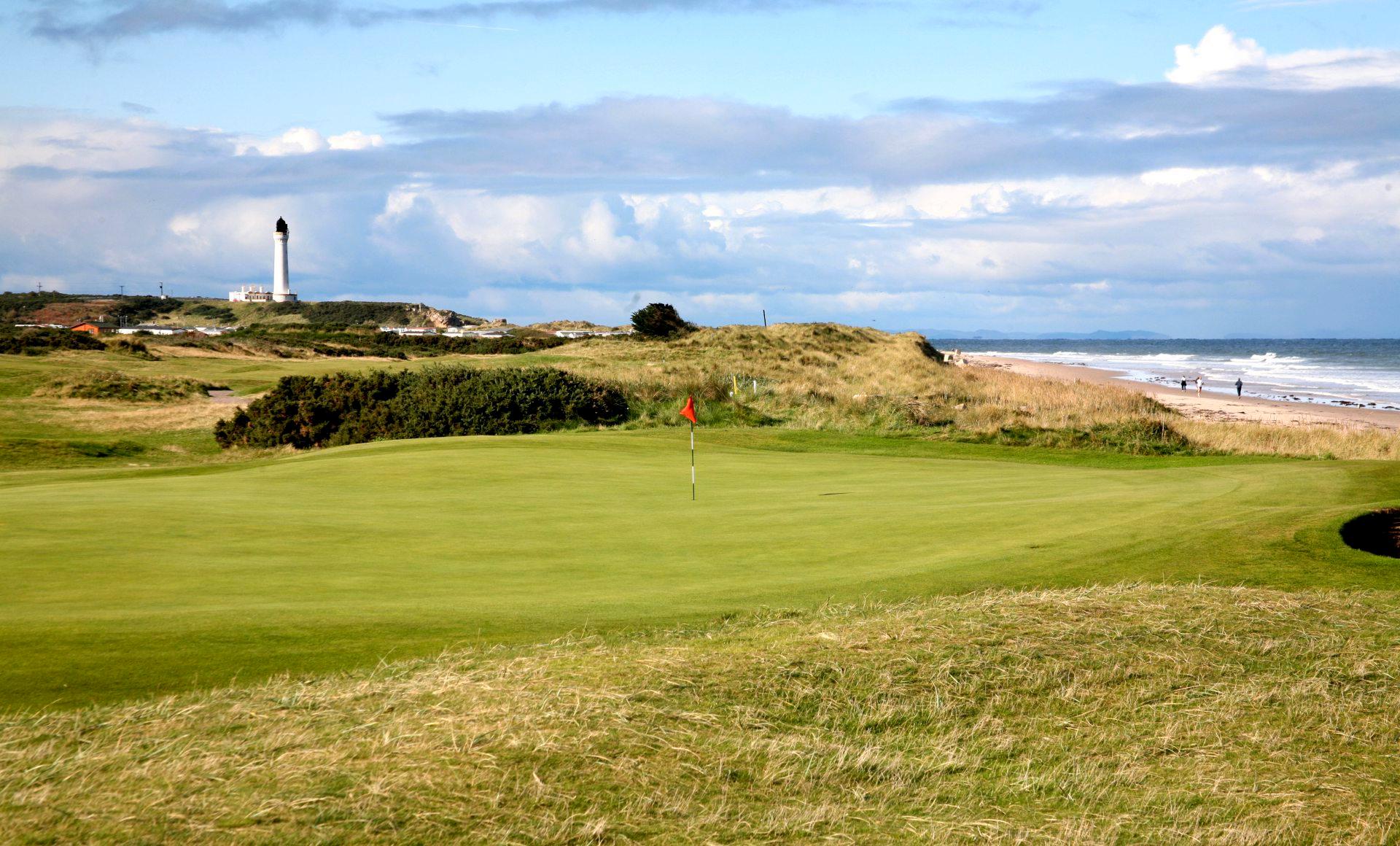 A green golf course with a lighthouse visible in the distance.