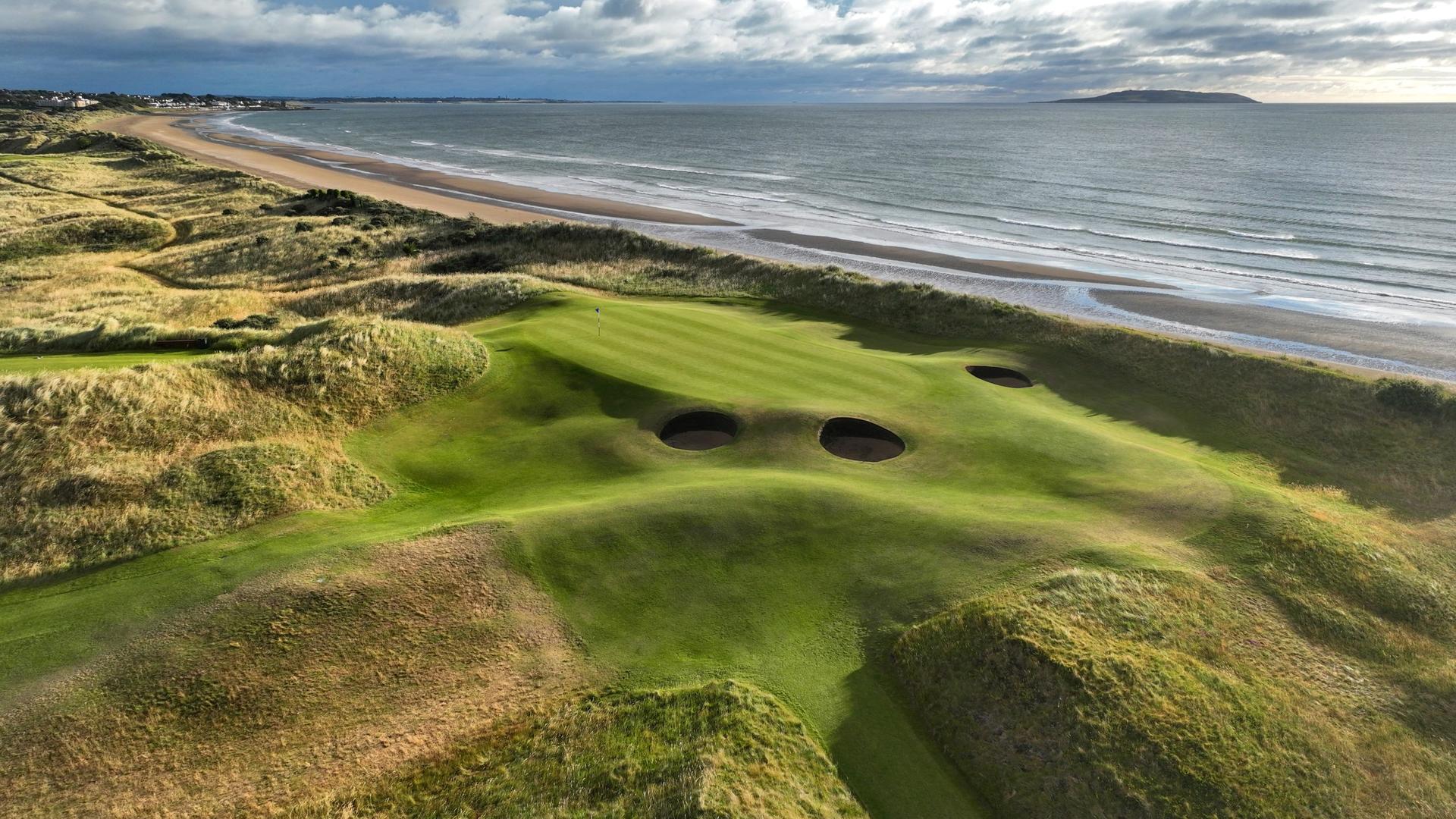A green with deep bunkers sits above a sweeping beach and ocean.