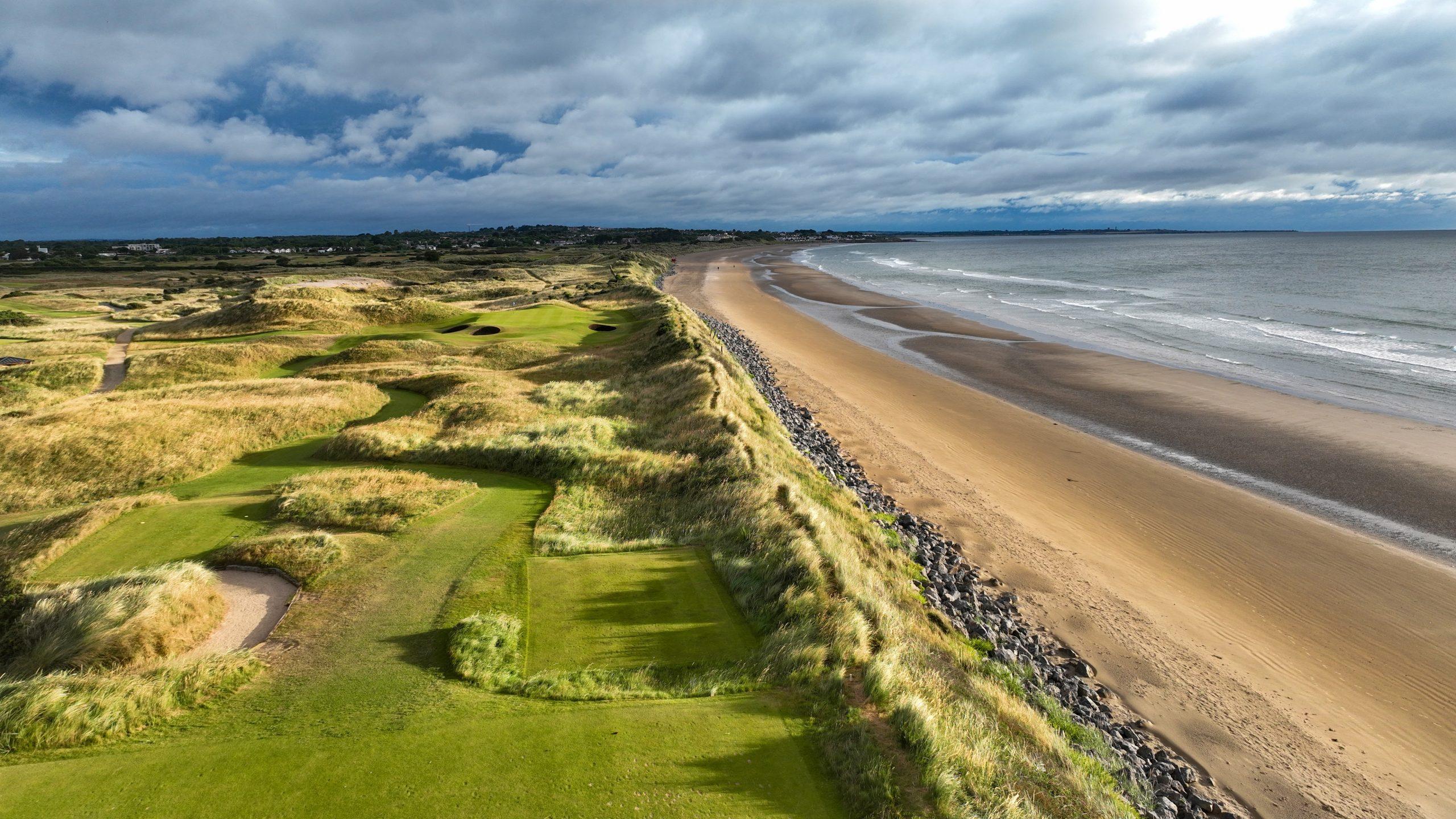 A coastal golf course runs alongside a sandy beach under cloudy skies.