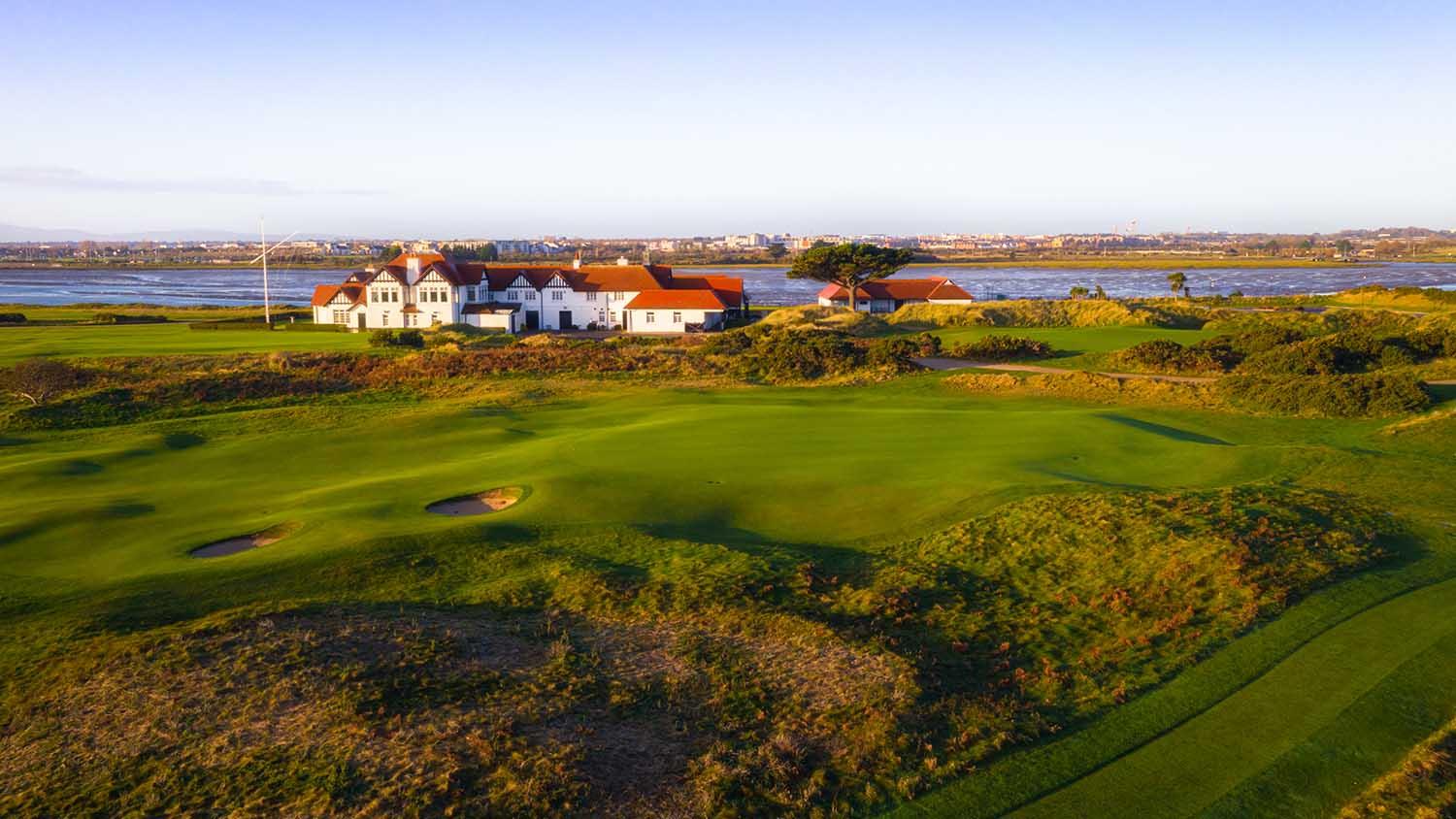 A clubhouse sits beside lush golf greens with water views in the distance.
