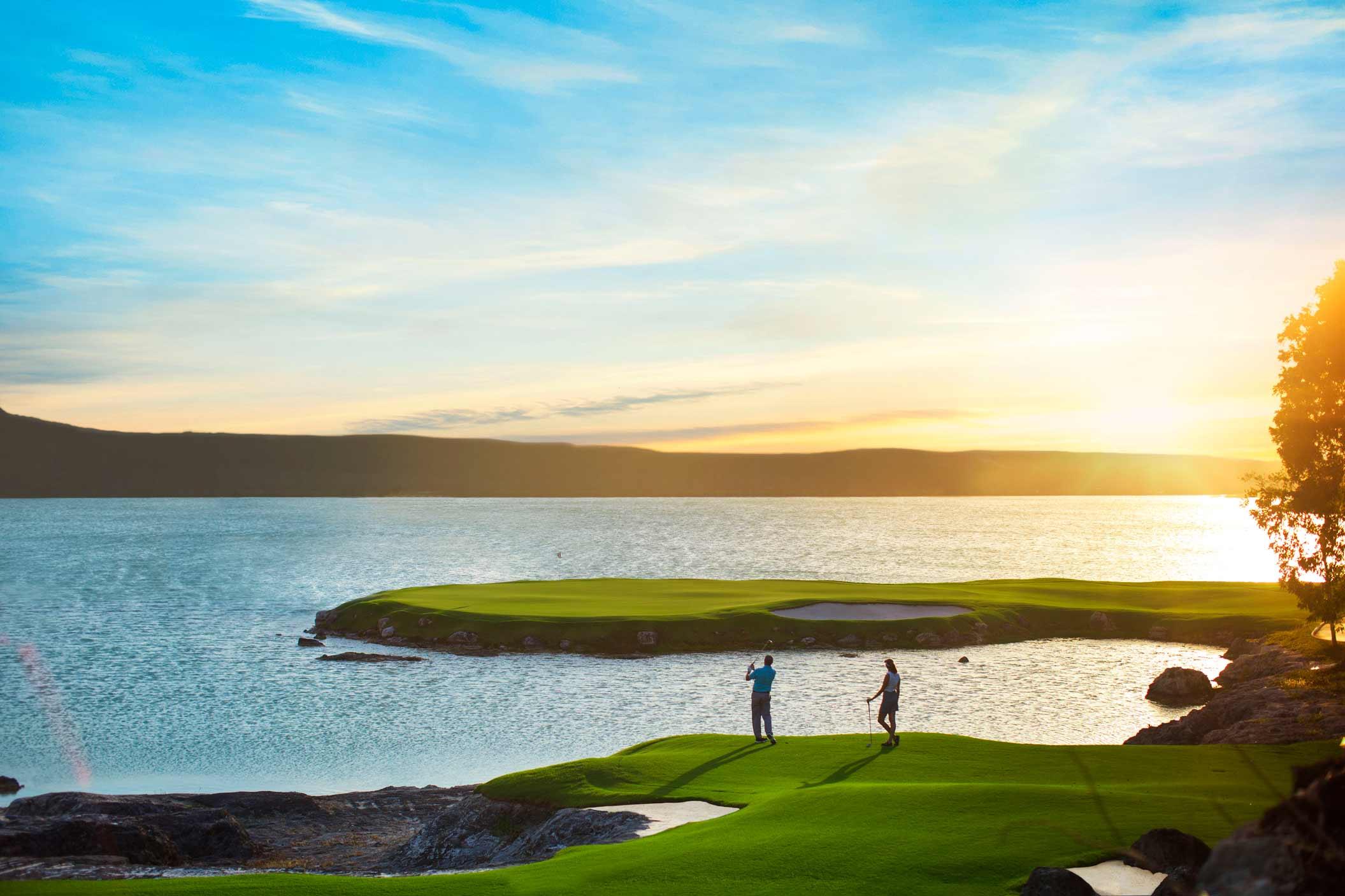 Two golfers play beside the water at sunset.