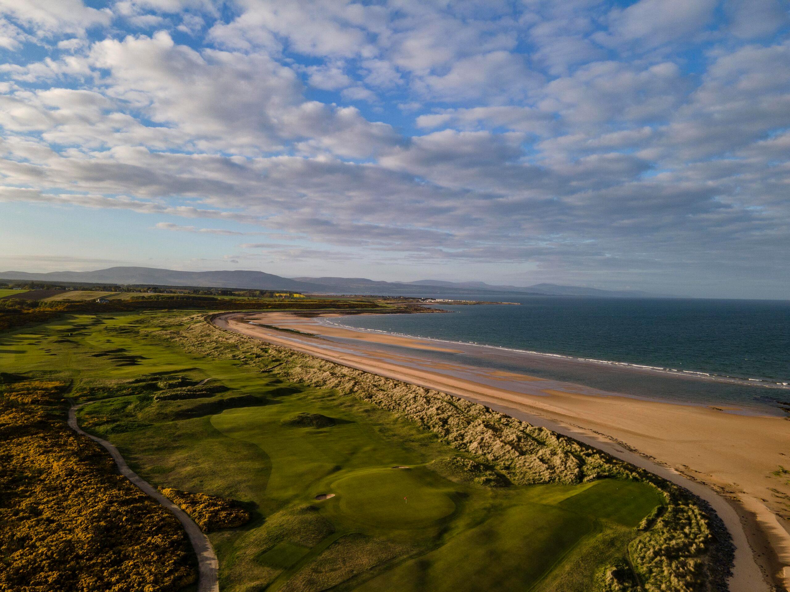 A golf course winds along a sandy coastline under blue skies.