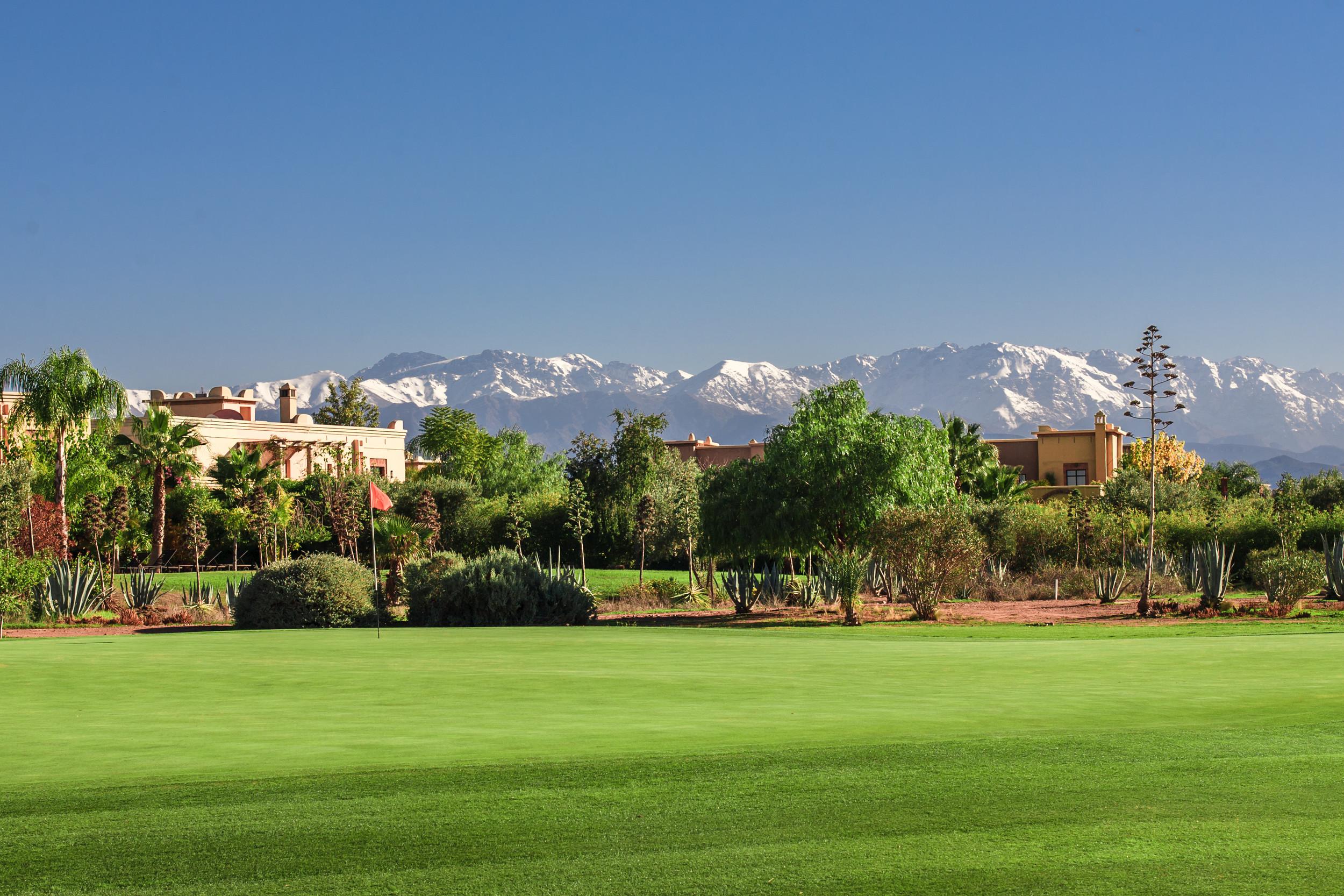 A lush green surrounded by trees and villas, with mountains towering in the background.
