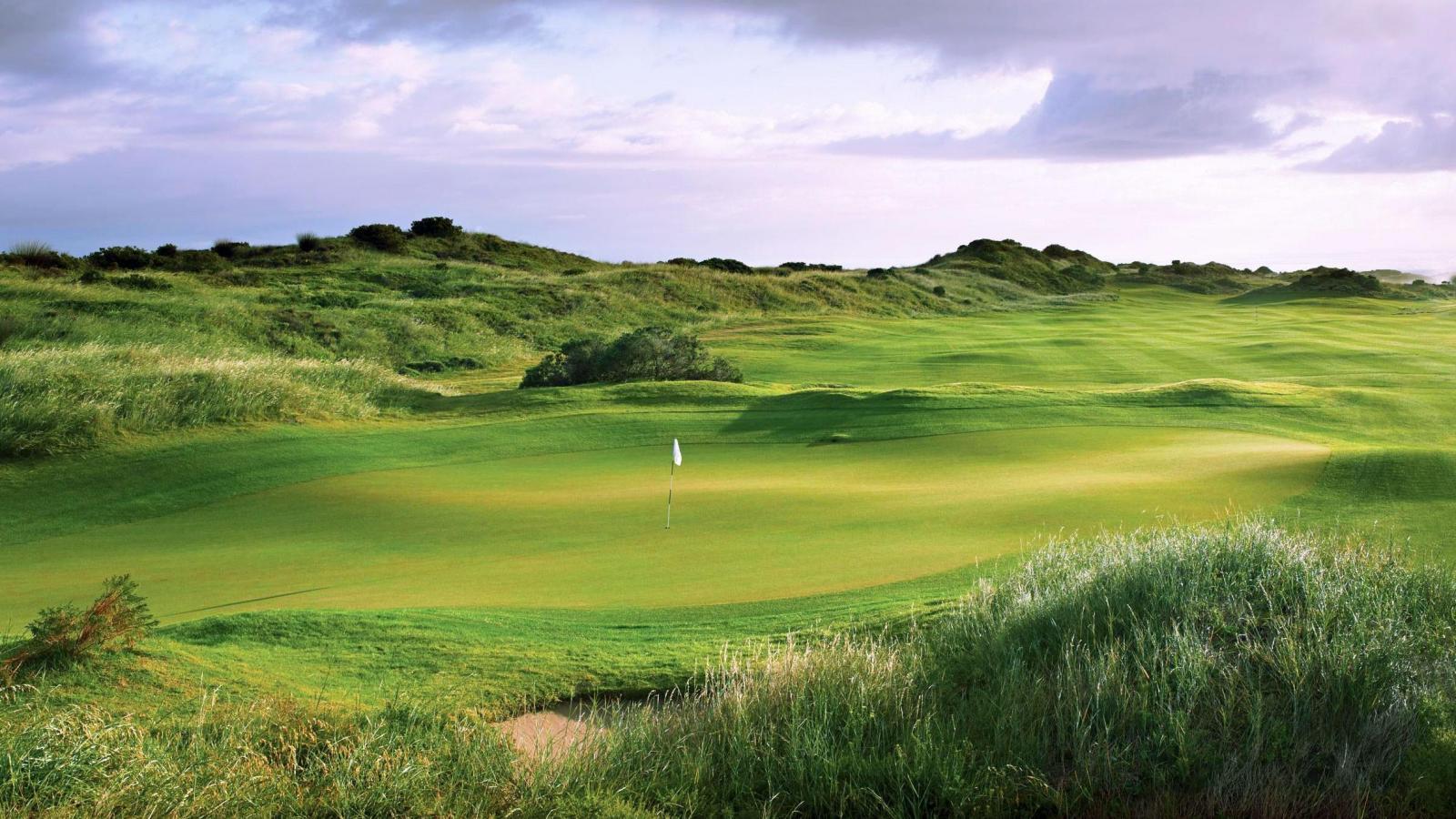 A lush green golf hole surrounded by rolling dunes and wild grass under a cloudy sky.