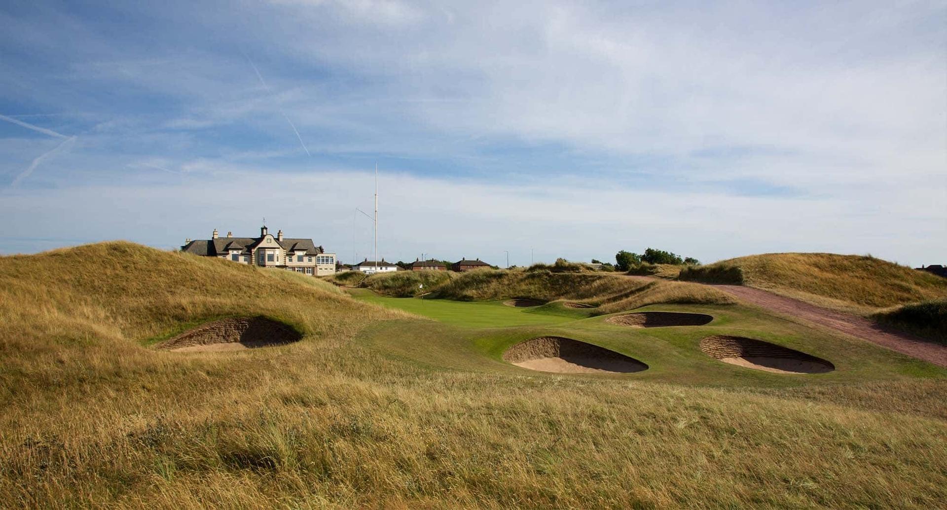 Multiple deep bunkers surround a green, with a clubhouse visible beyond the golden grassy dunes.