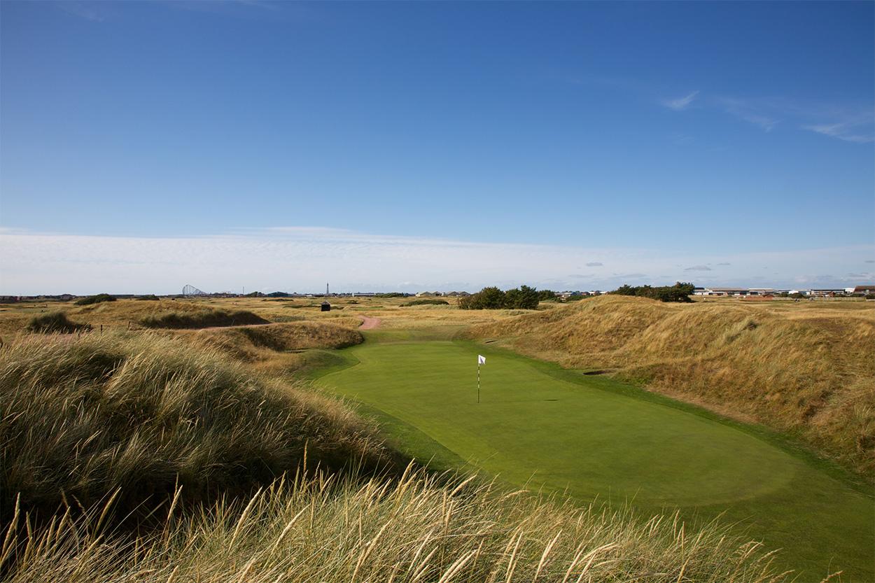 A putting green nestled between high grassy dunes, with a rollercoaster visible faintly in the background.