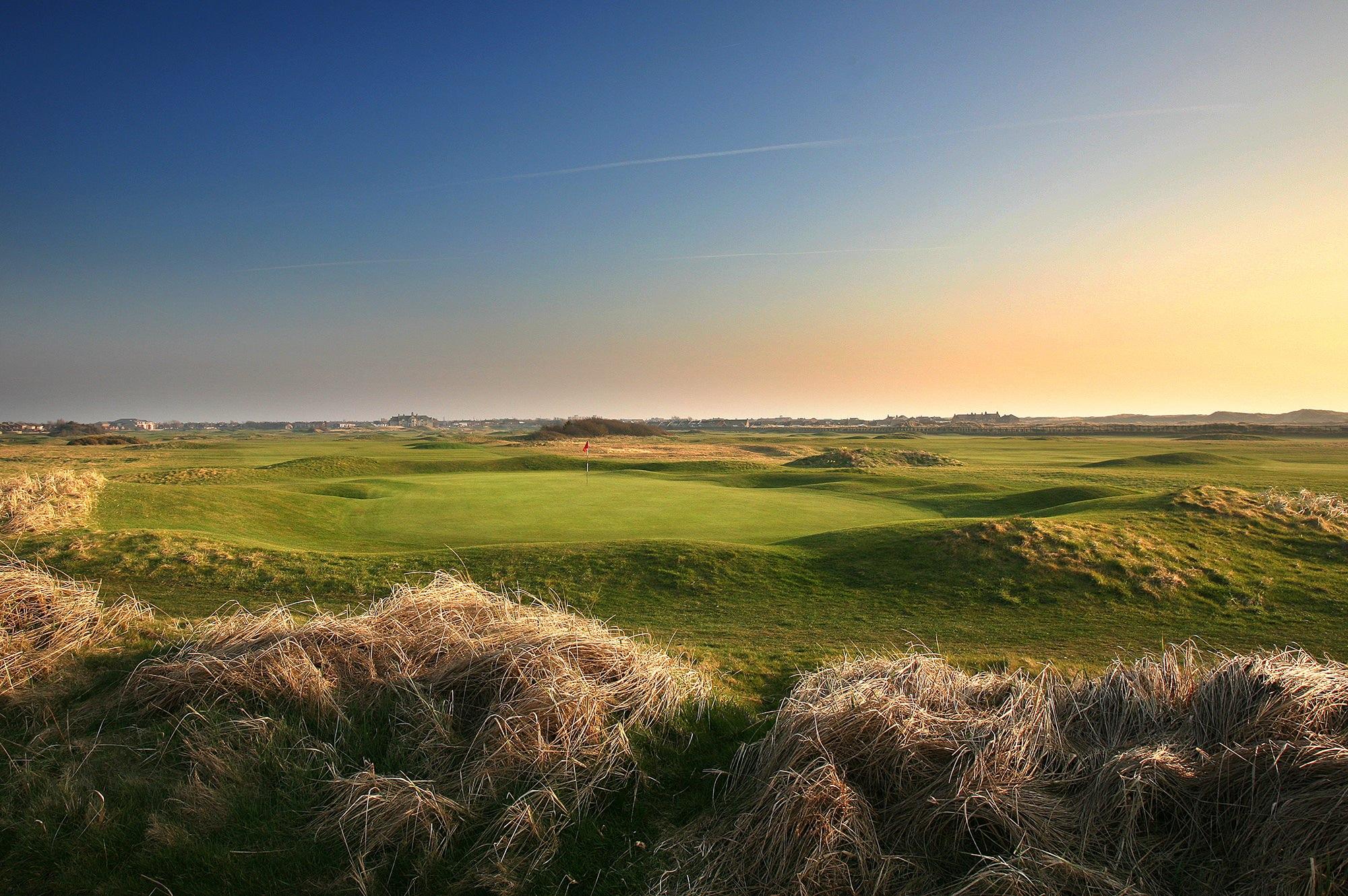 A wide view of a flat, open golf green surrounded by tall golden grass under a clear evening sky.