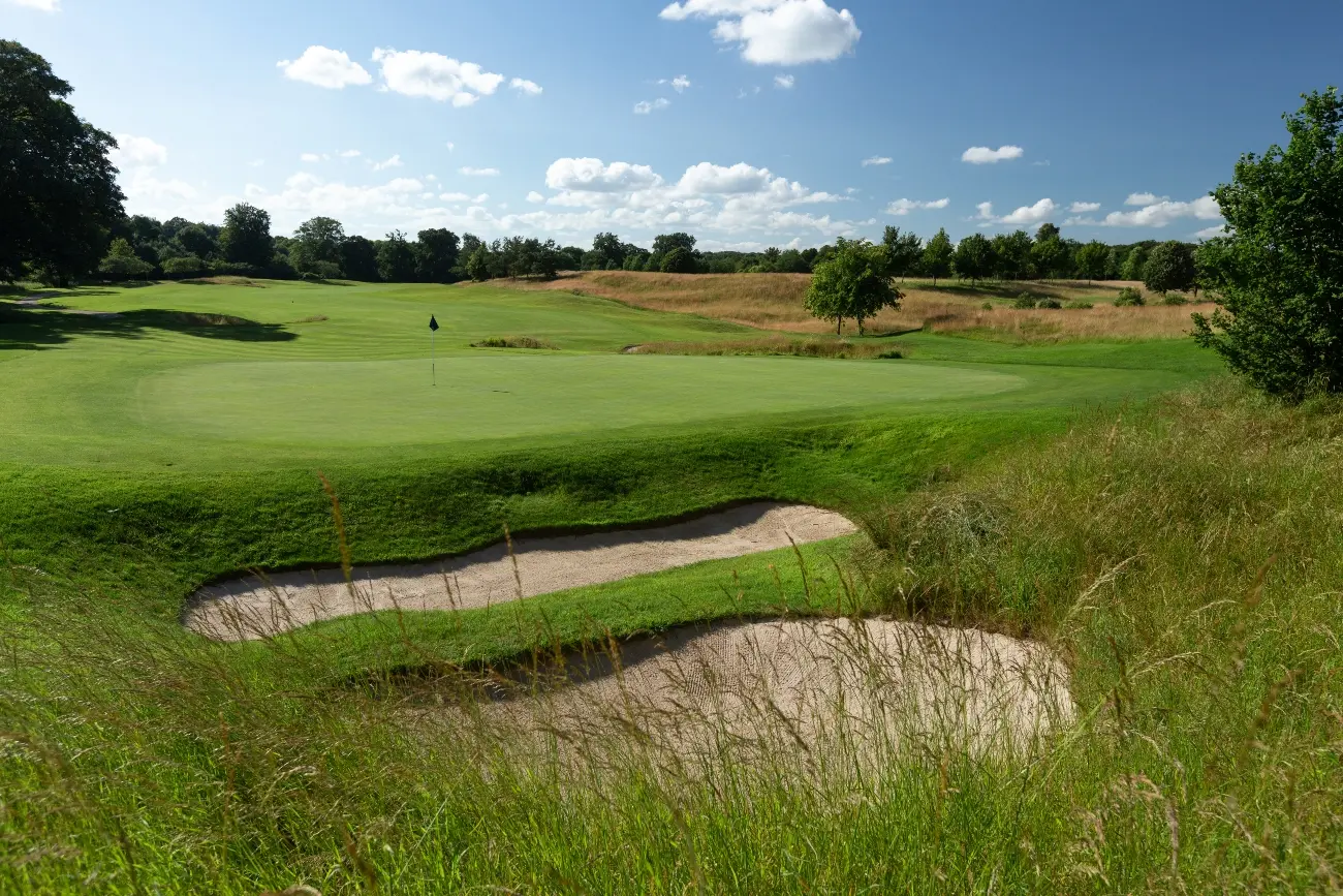 A lush green fairway with deep bunkers framed by tall grass under a bright blue sky.