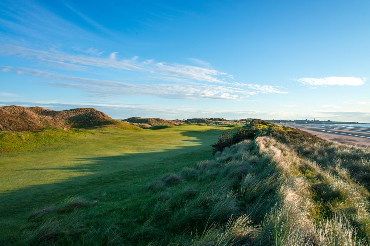 A pristine fairway nestled between tall dunes, with sweeping views of the beach and ocean under a bright blue sky.
