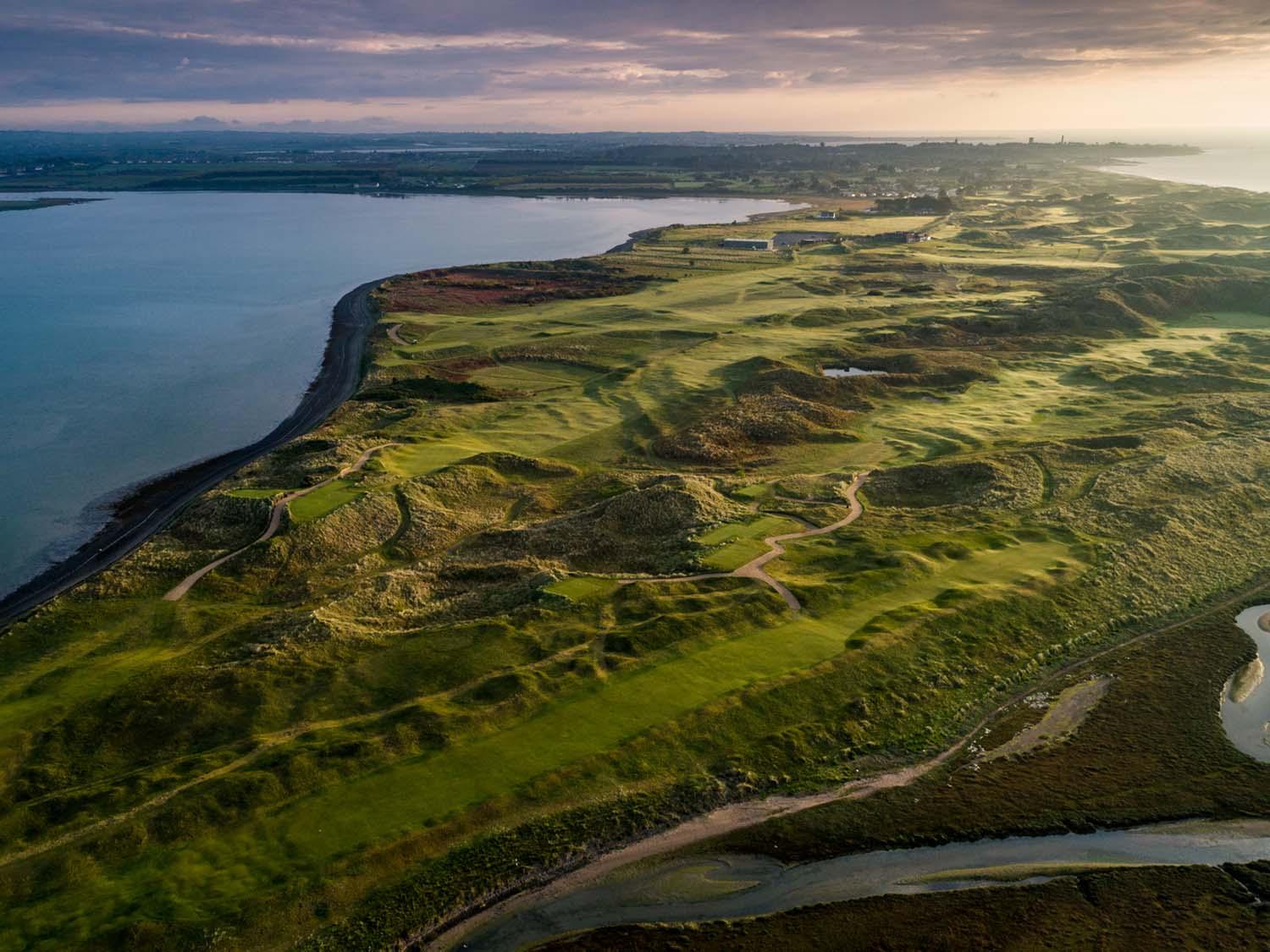 A dramatic aerial view of a golf course surrounded by rugged dunes, with winding paths and a wide body of water along its edge.