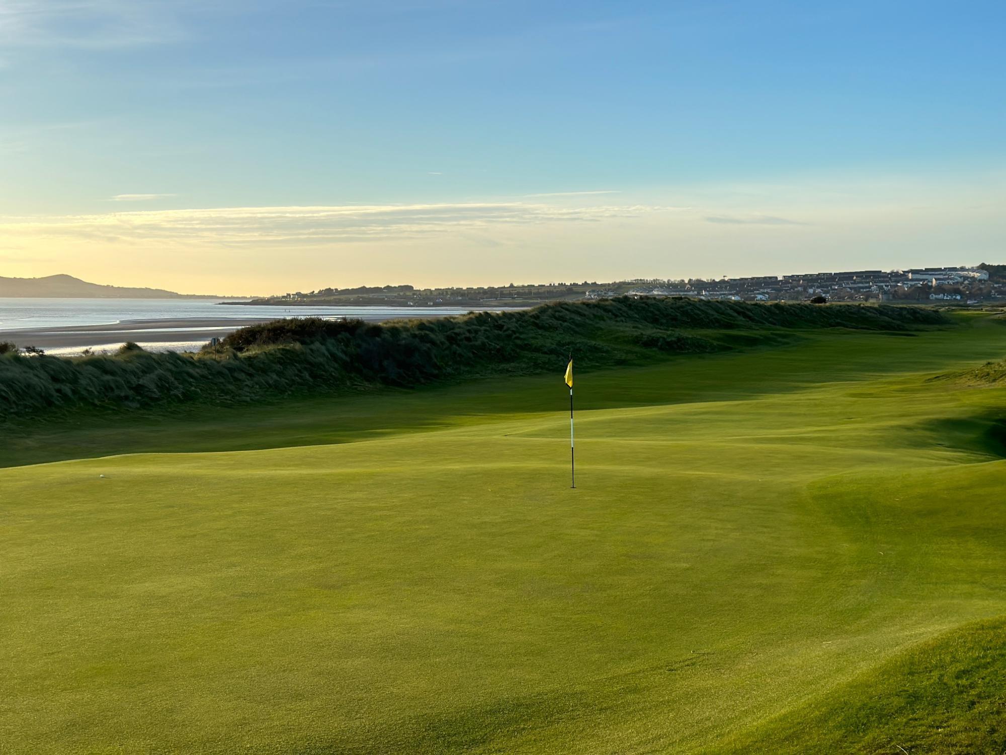 A beautifully maintained green with a golf flag, bordered by dunes and overlooking a calm ocean with a coastal town in the distance.
