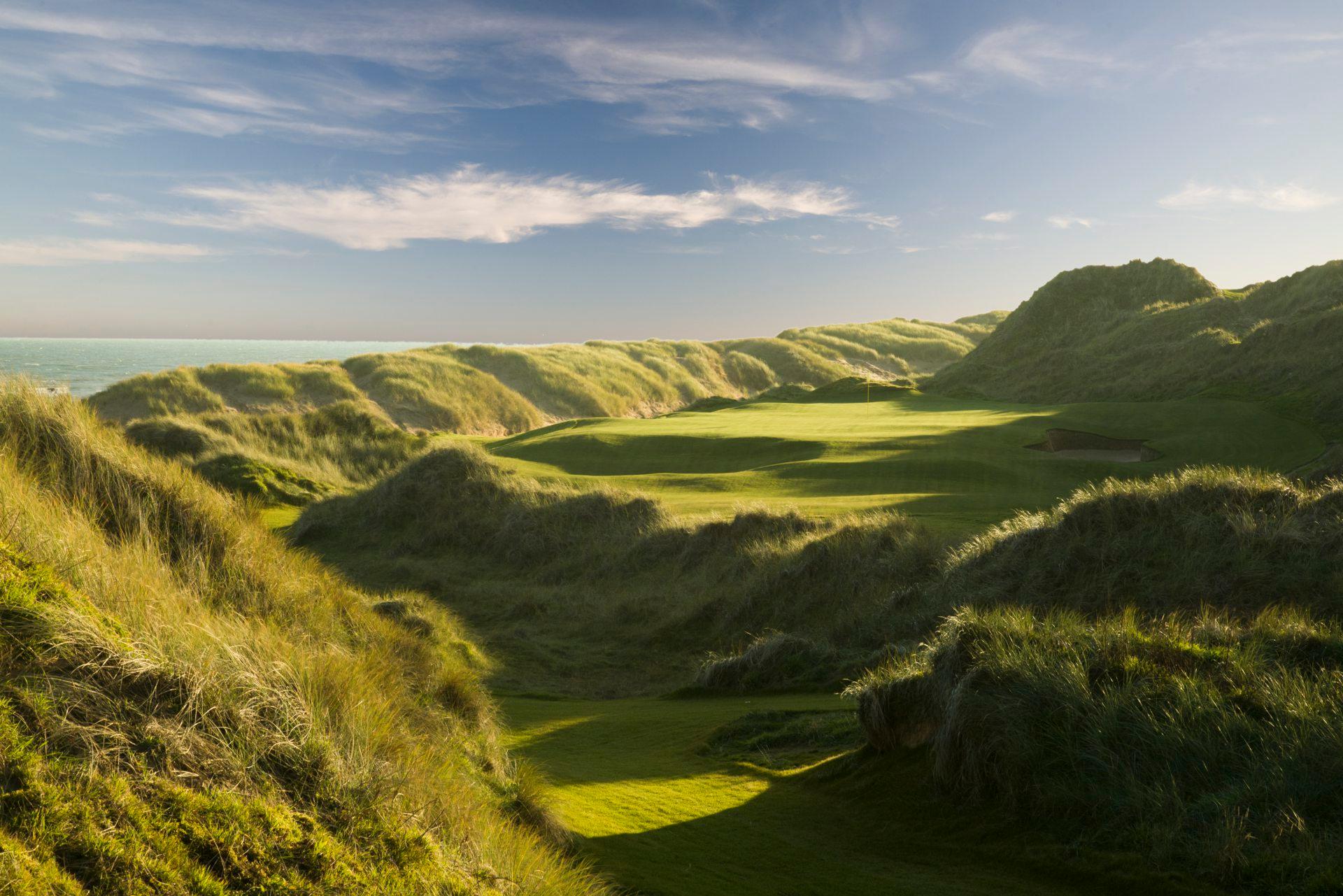 A green fairway surrounded by towering dunes near the ocean.