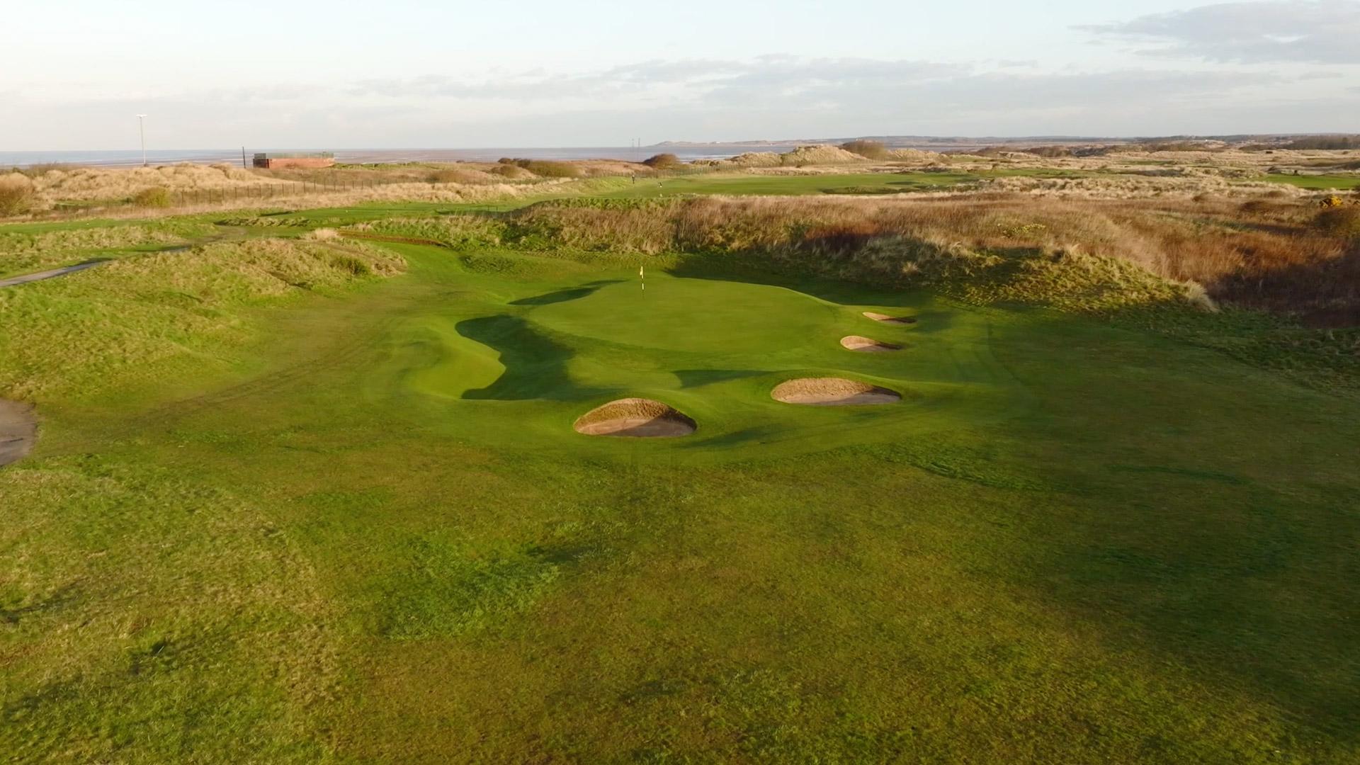 An elevated green protected by several pot bunkers, framed by rugged dunes.