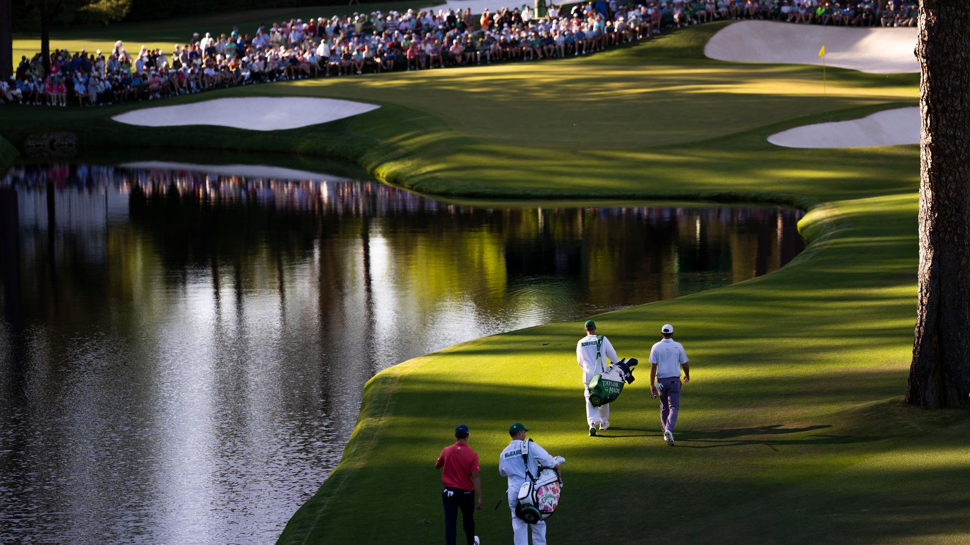 Scheffler and Hojgaard walking with their caddies along a winding fairway with a large water hazard on the left