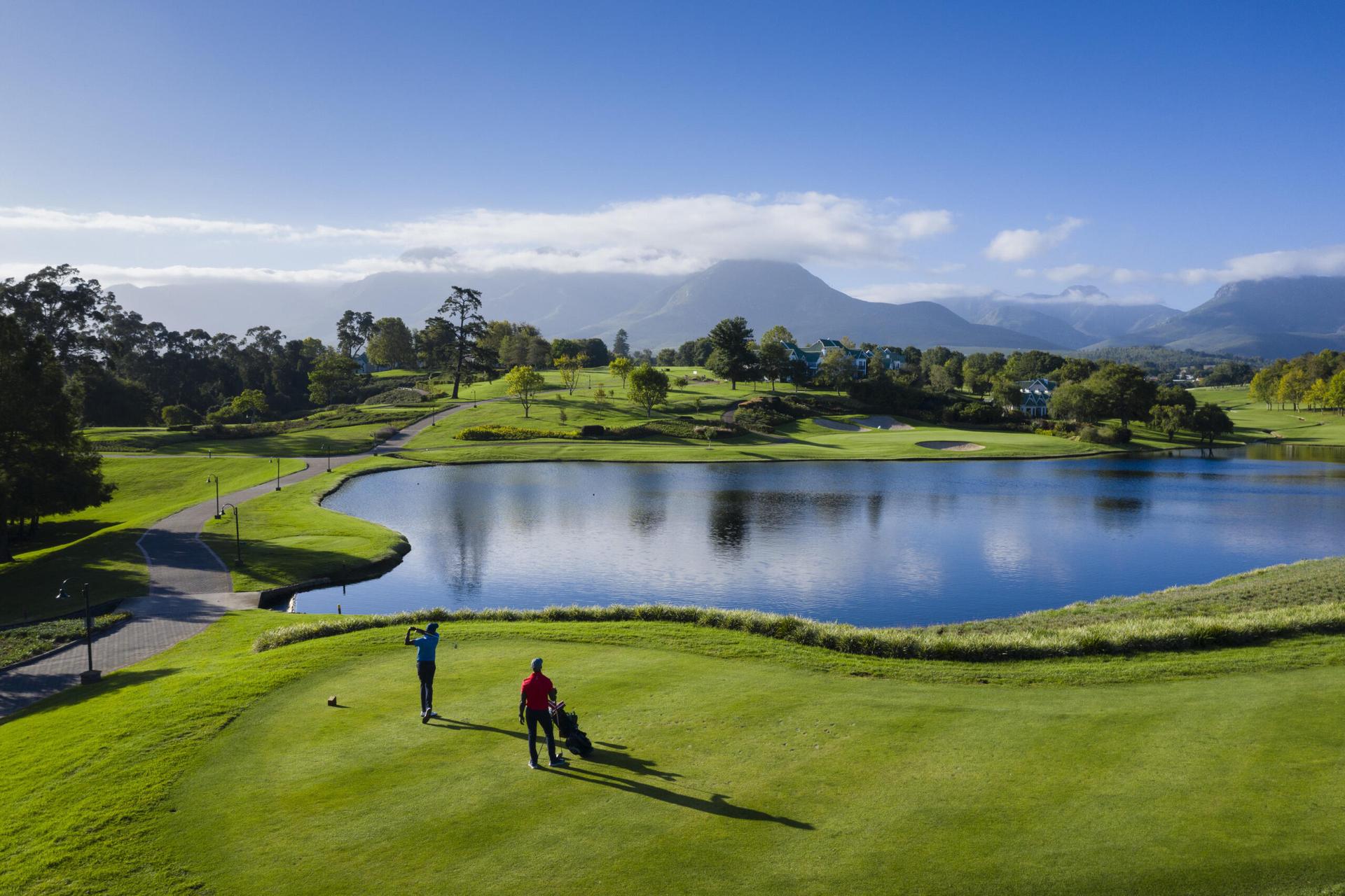 Two players on a tee box at Fancourt Links over a large water hazard with mountains in the background