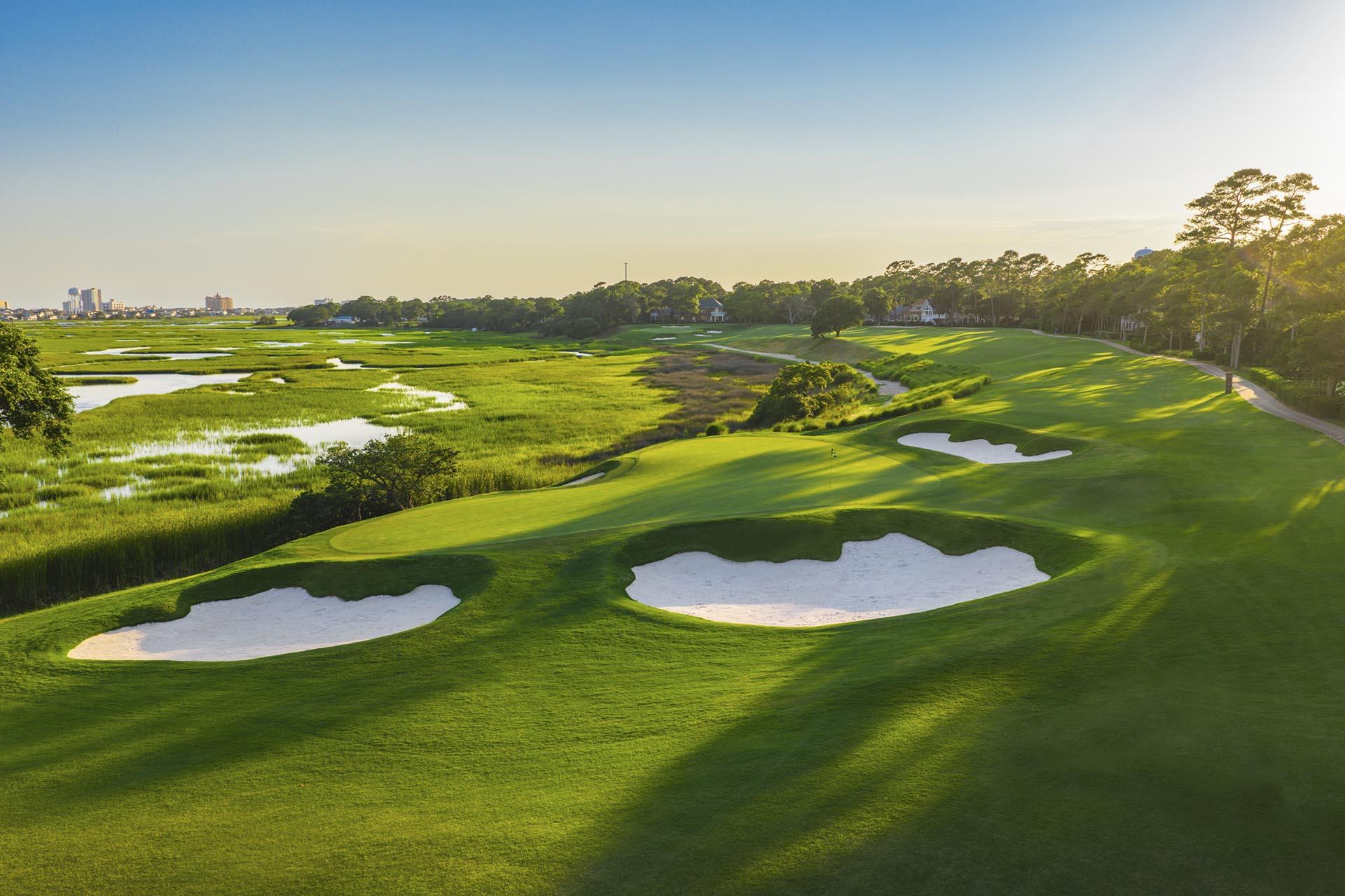 Narrow fairway winding around marshland to a small, sloping green with bunkers around it