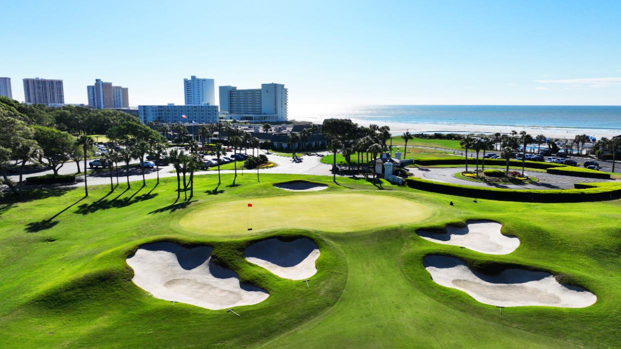 A smooth green surrounded by sand bunkers with city views in the distance
