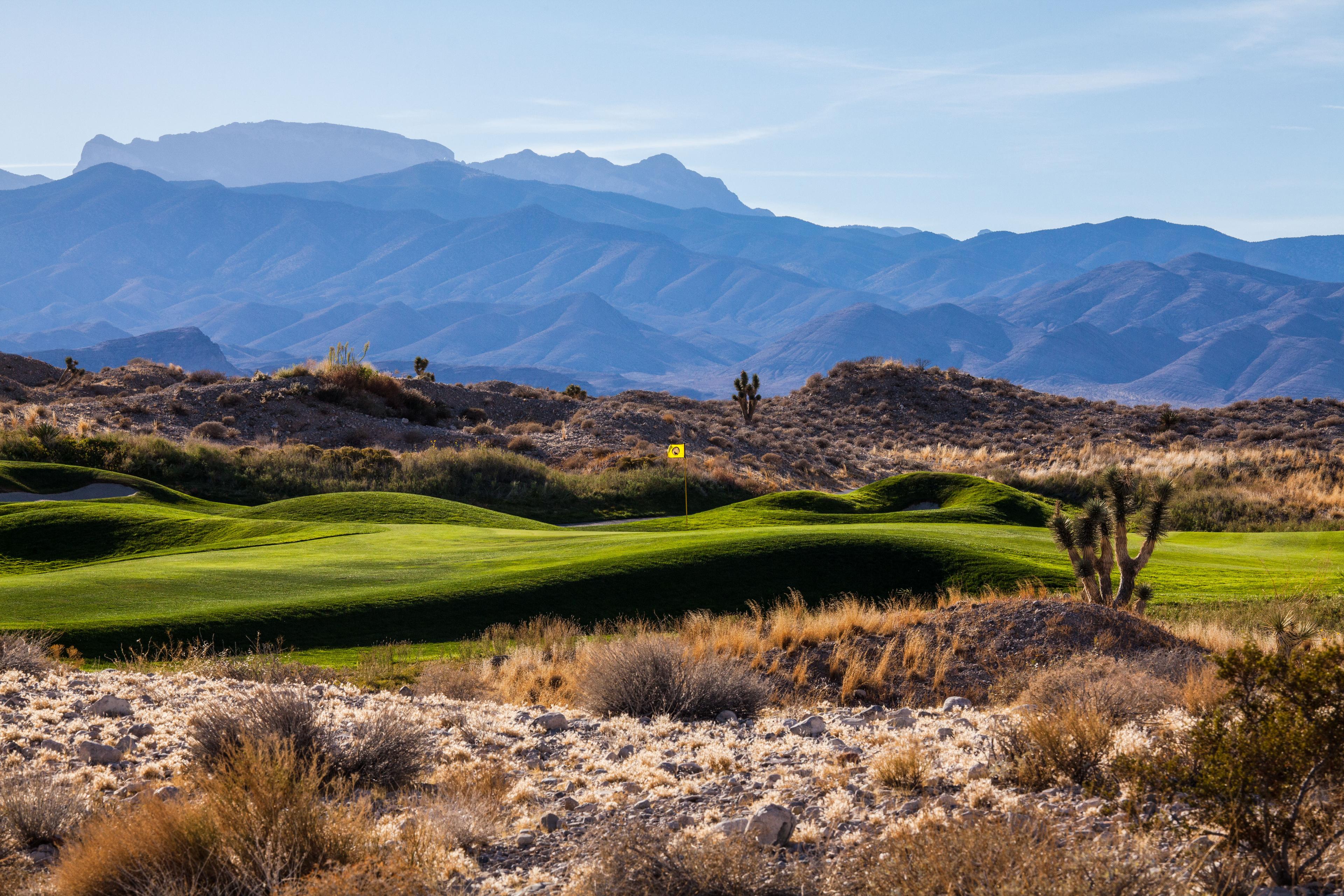 A manicured green surrounded by rolling dunes with mountains in the distance