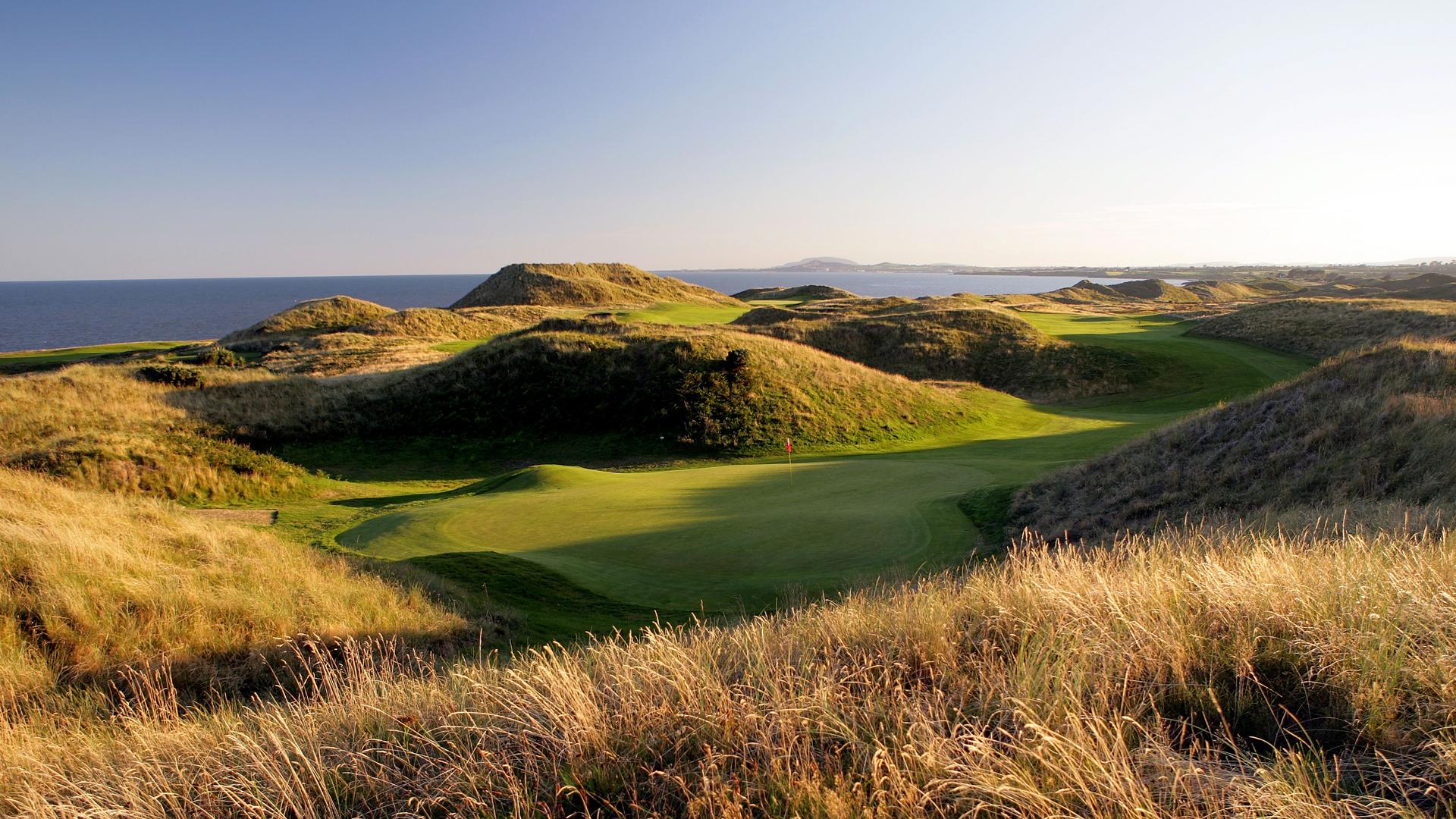 Overhead view of a winding fairway leading to smooth green with ocean views in the background