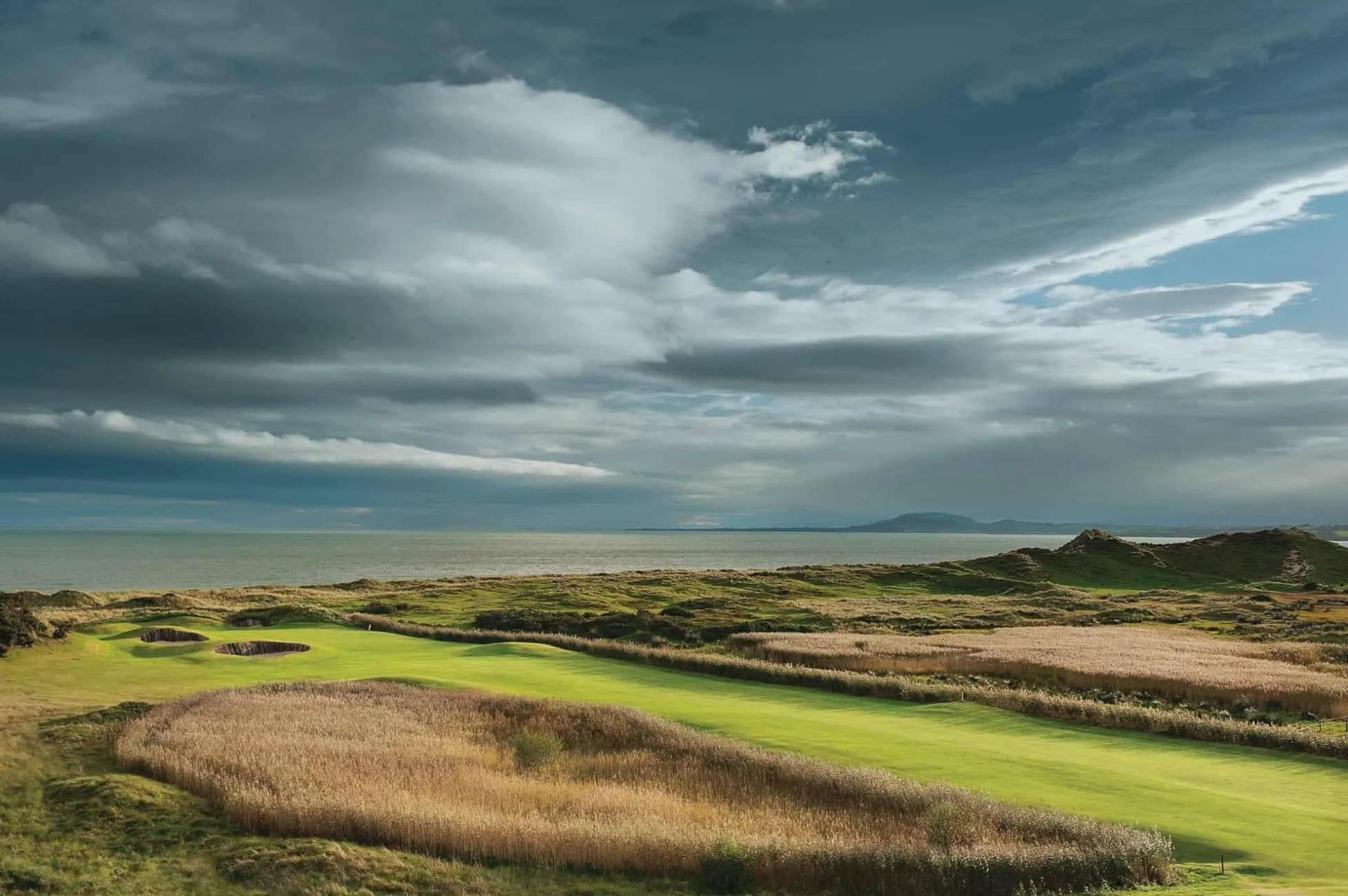 A straight fairway leading to smooth green surrounded by sand bunkers with costal views in the background