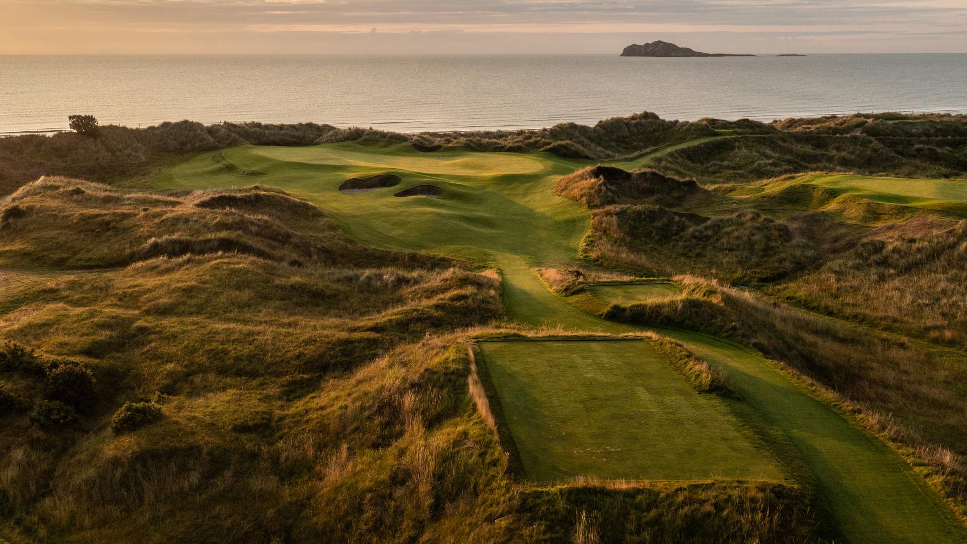 A tee box facing towards the coast next to a smooth green surrounded by sand bunkers and rolling dunes