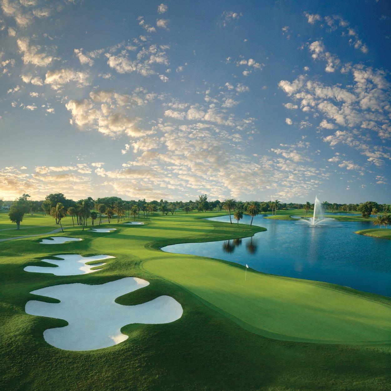 A lush golf course with pristine white sand bunkers and a large water feature with a fountain under a beautiful cloud-filled sky.