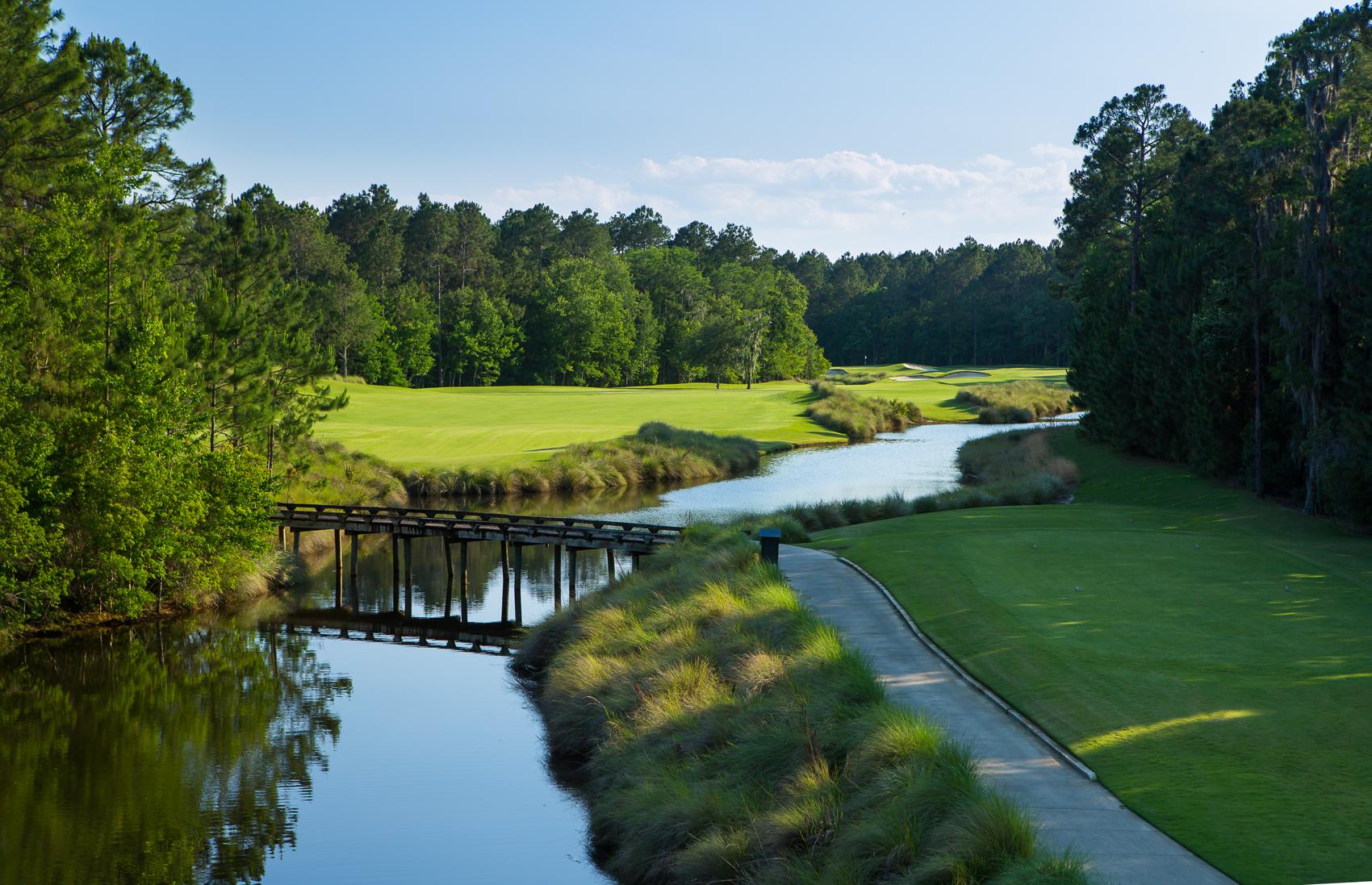 A narrow river winds through the golf course, with a wooden bridge connecting the fairways.