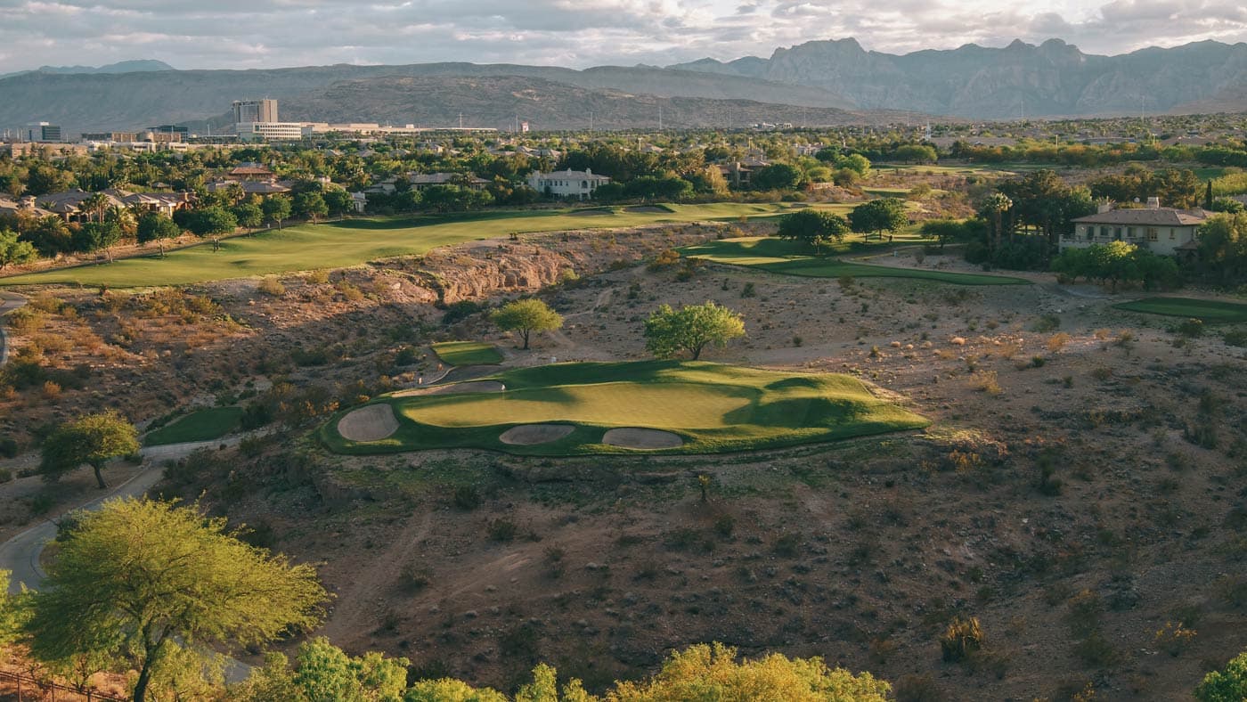 Birdseye view of an island like green littered with sand bunkers being surrounded by a rocky rough with mountains in the distance