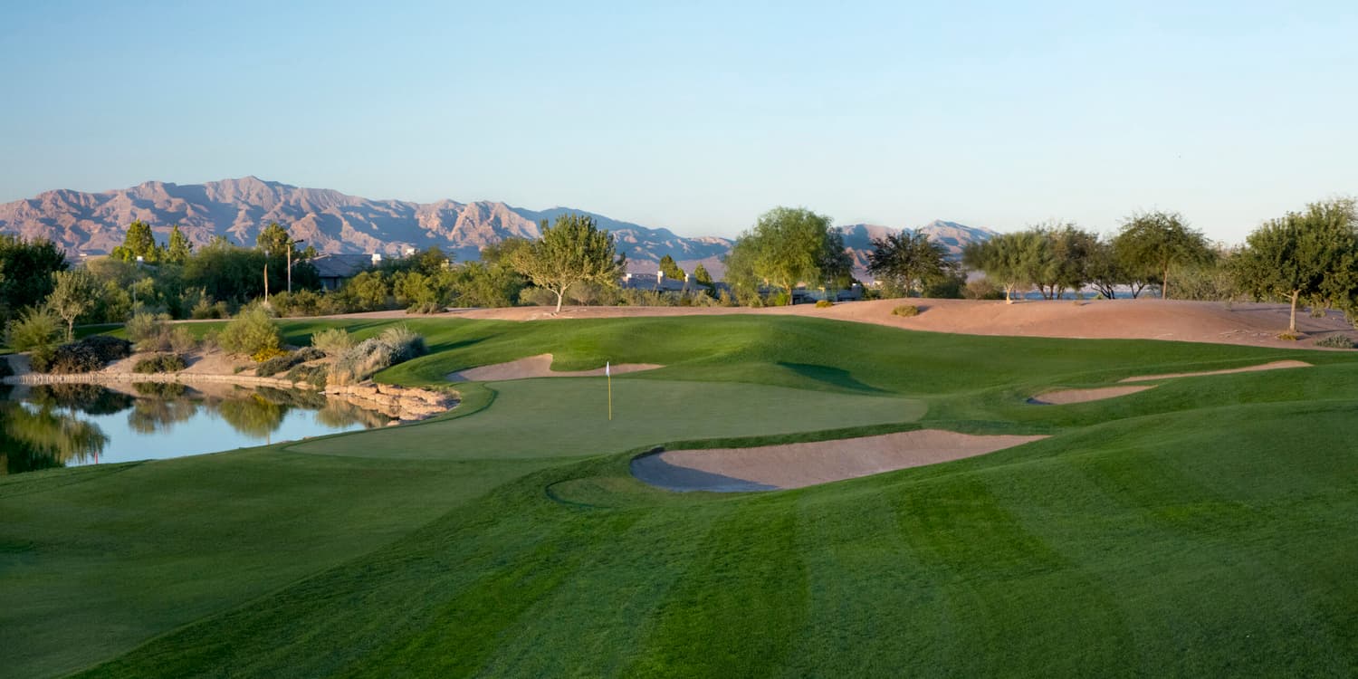 A smooth green littered with sand bunkers between rolling dunes and a water hazard reflecting the sky