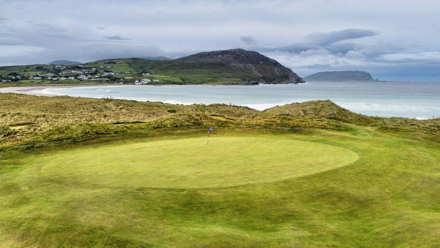 This shows a close up of a hole at the course with manicured greens an ocean background with mountains towering over