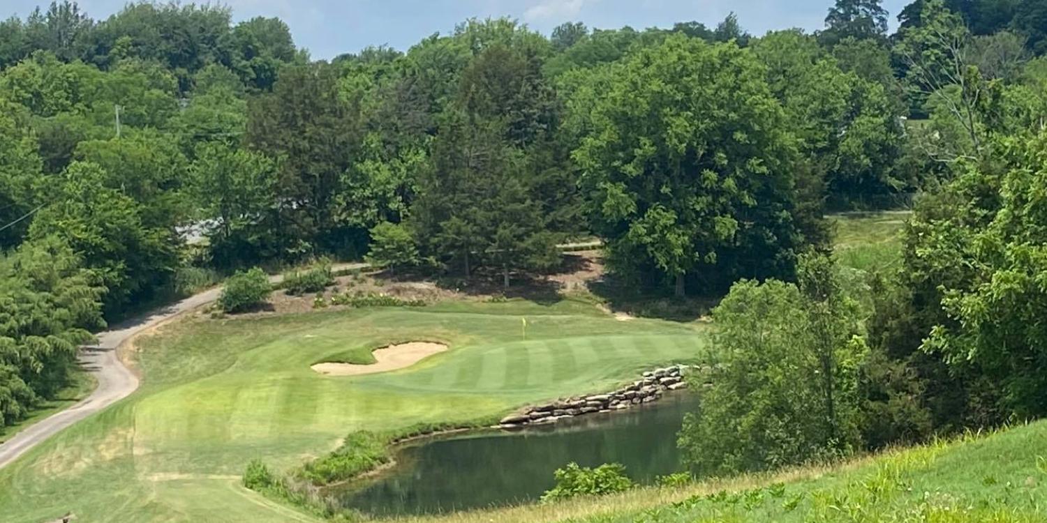 A scenic golf hole is surrounded by lush trees, a small pond, and a stone-lined water feature near the green.