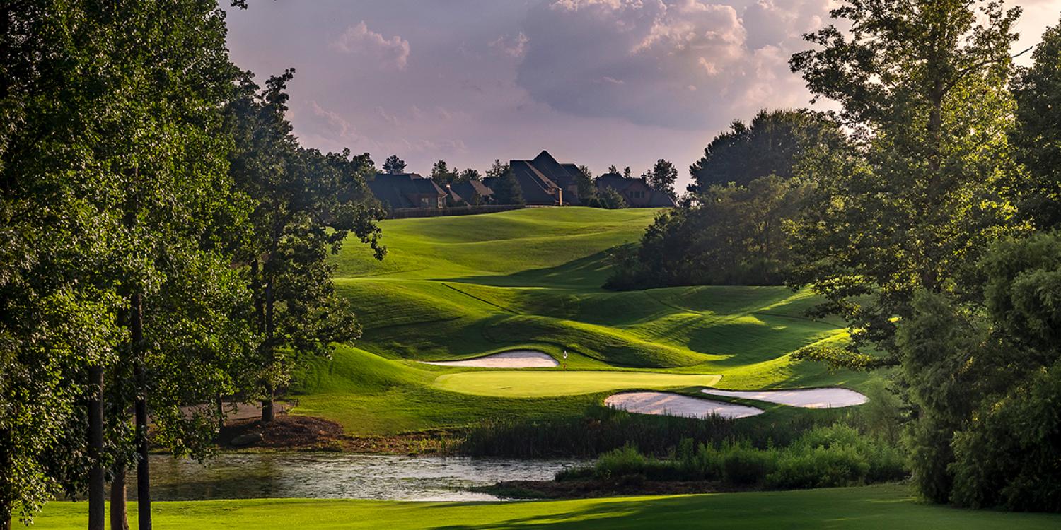 A pristine golf course is framed by tall trees and natural water features, with bunkers adding challenge to the green.