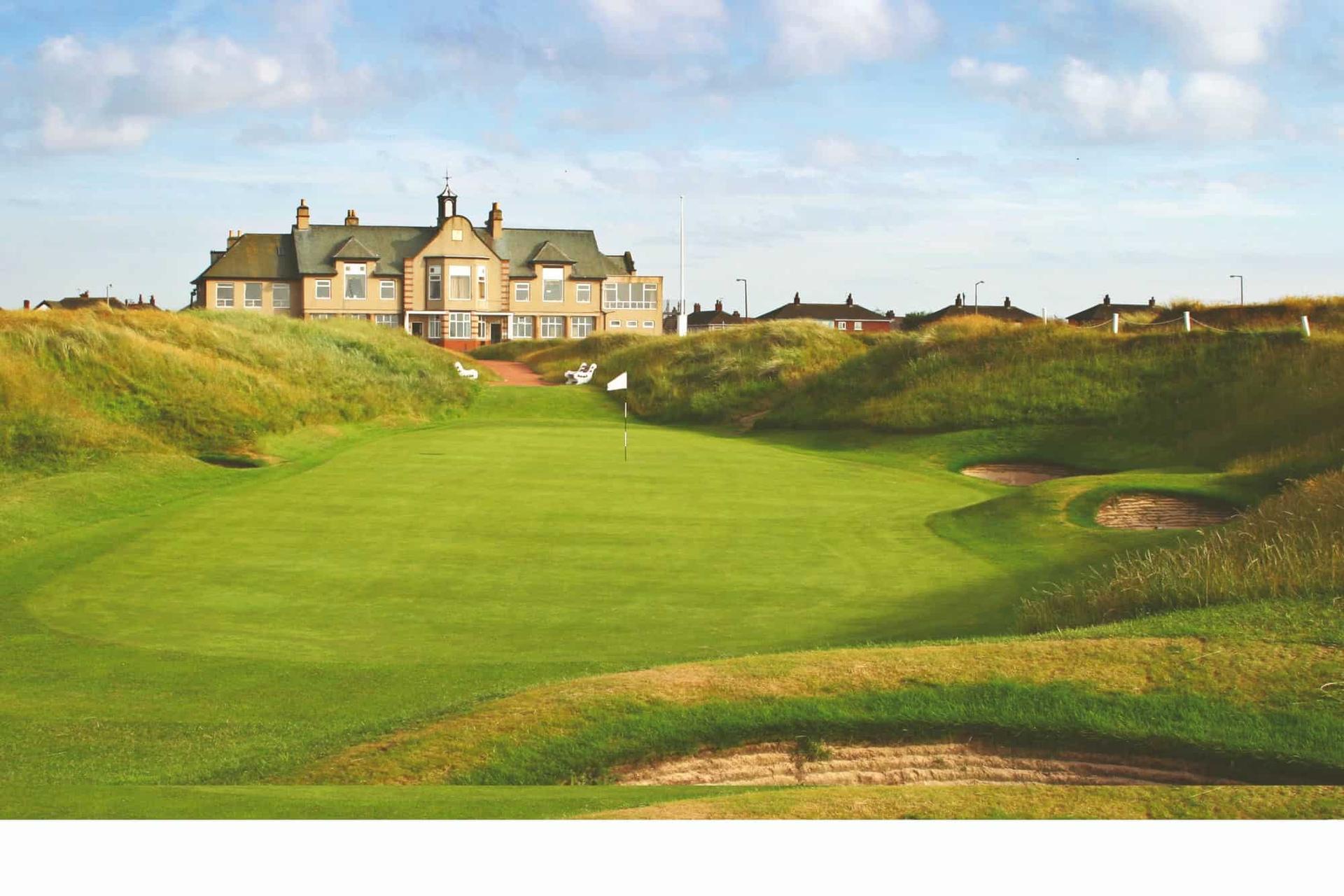 A golf green with bunkers, framed by grassy dunes, with a clubhouse standing prominently in the background.