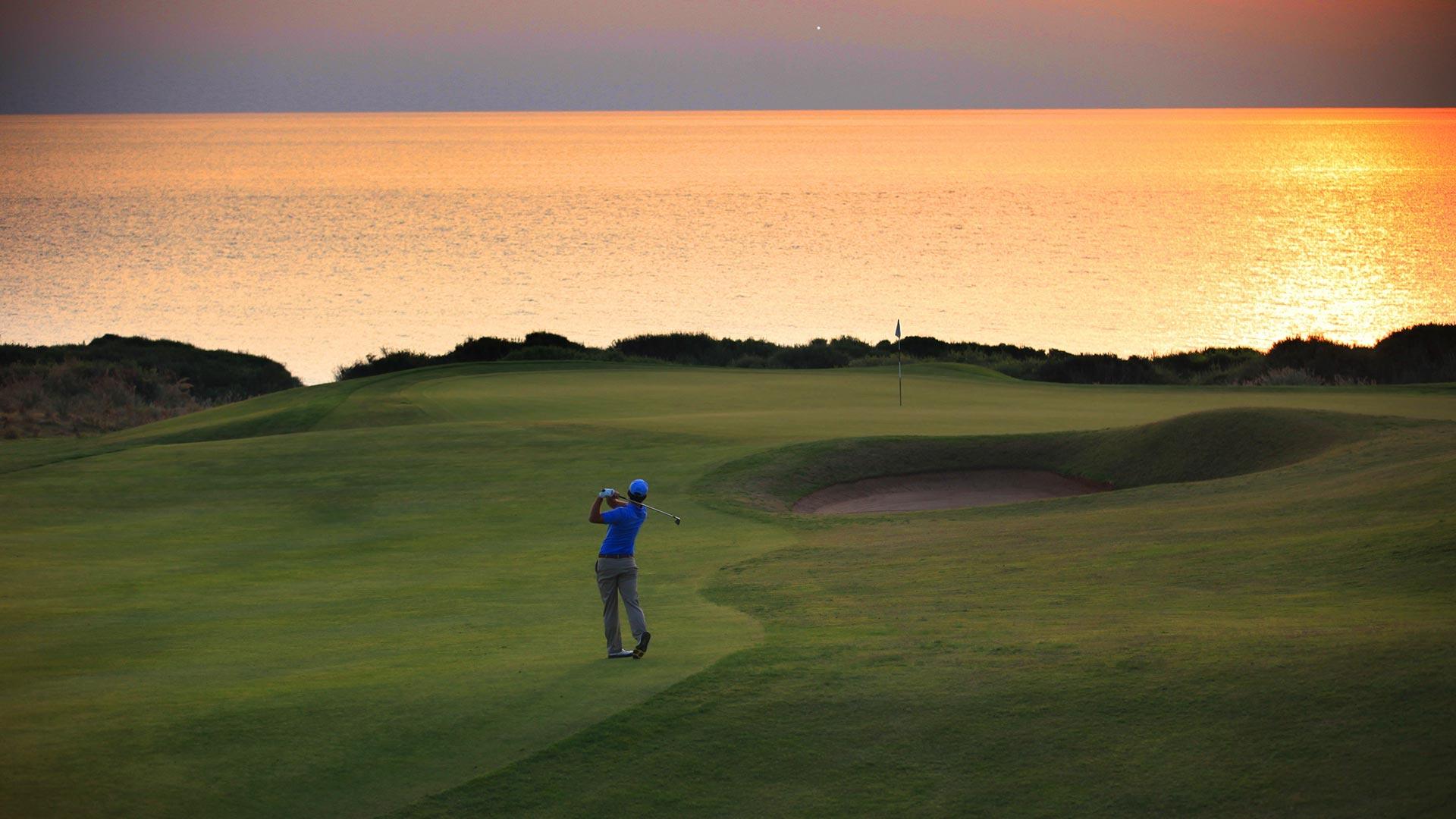 A golfer swings toward a seaside green under a glowing orange sunset.