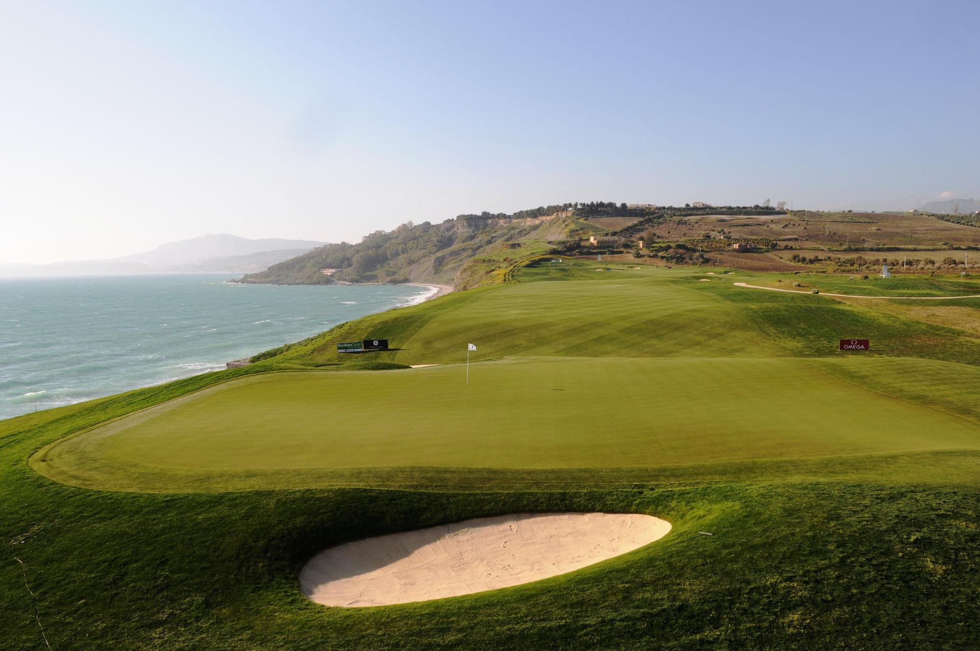 A wide fairway leading to a well maintained green with coastal views under clear skies