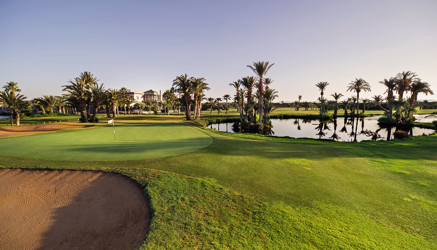 A luxurious green with palm trees reflecting in a tranquil pond at sunset.