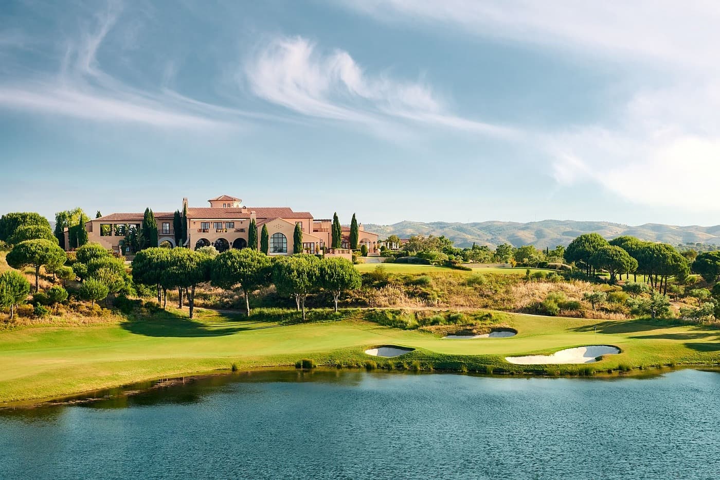 A clubhouse building overlooking a smooth green surrounded by sand bunkers next to a water hazard
