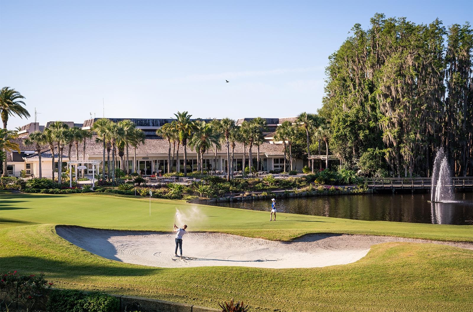 Golfer swinging out of a sand bunker onto a well maintained green next to a water hazard