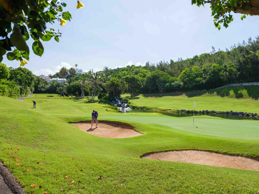 A golfer plays from a bunker on a lush, tree-lined course with a cascading waterfall near the green.