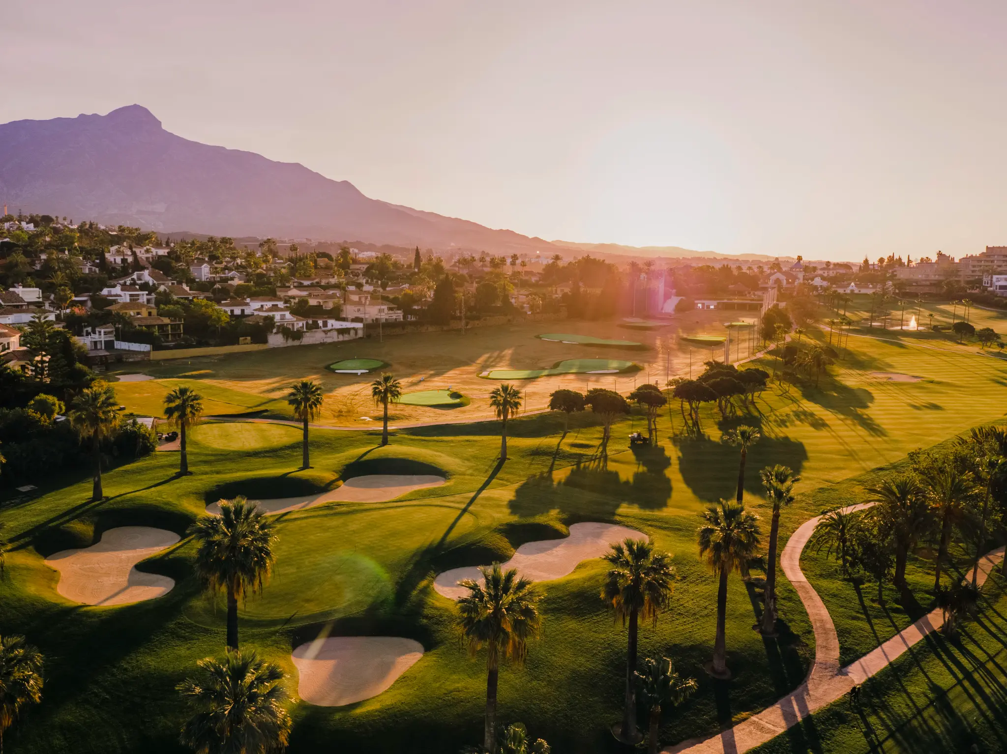 Overhead view of the sun setting over a well maintained fairway leading to a smooth green surrounded by sand bunkers