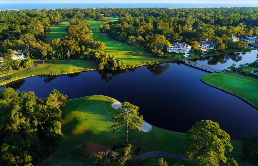 A scenic aerial view of a golf course with lush fairways surrounded by water and nearby houses, creating a serene coastal setting.