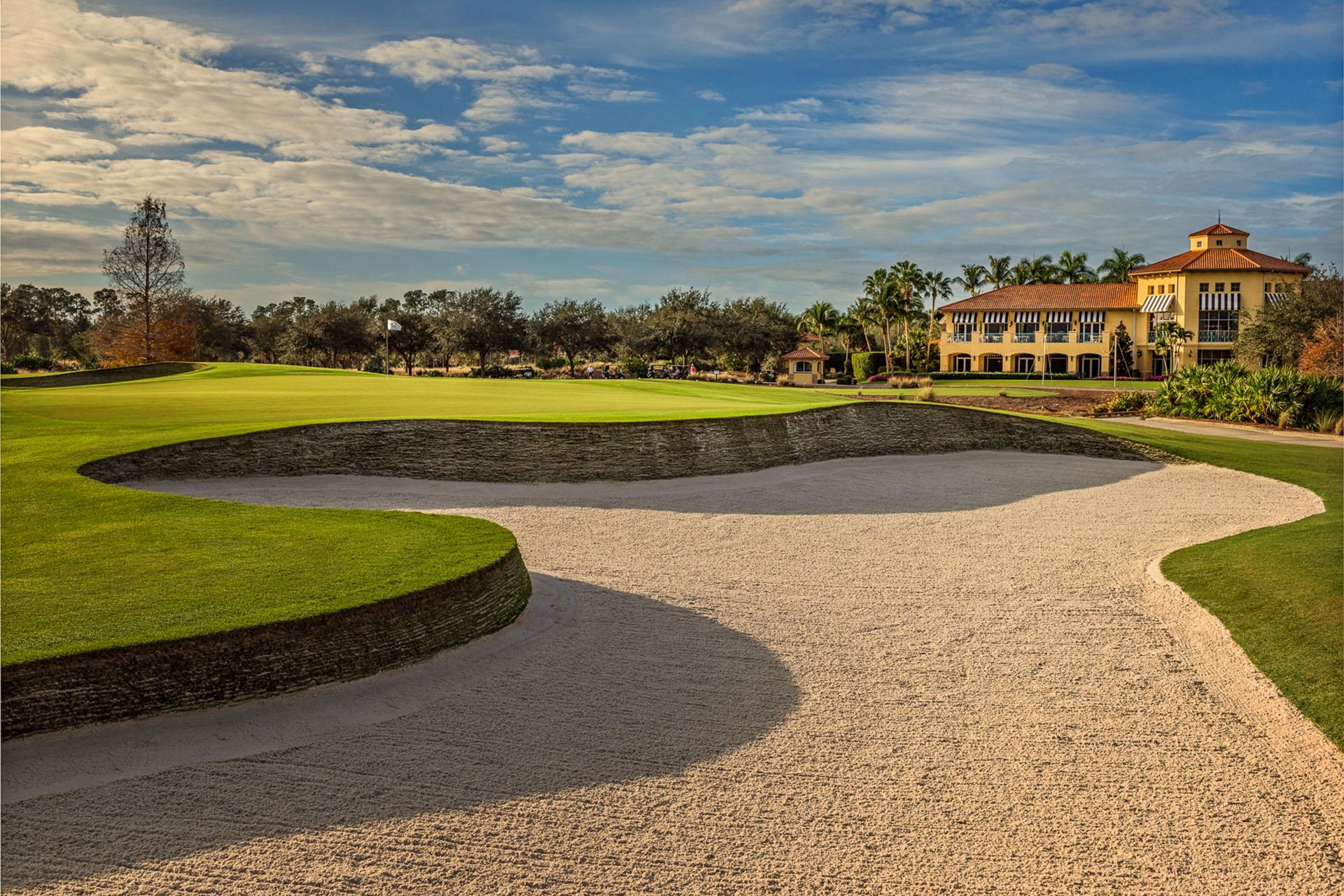 A pristine bunker sits near the green with a clubhouse in the background.