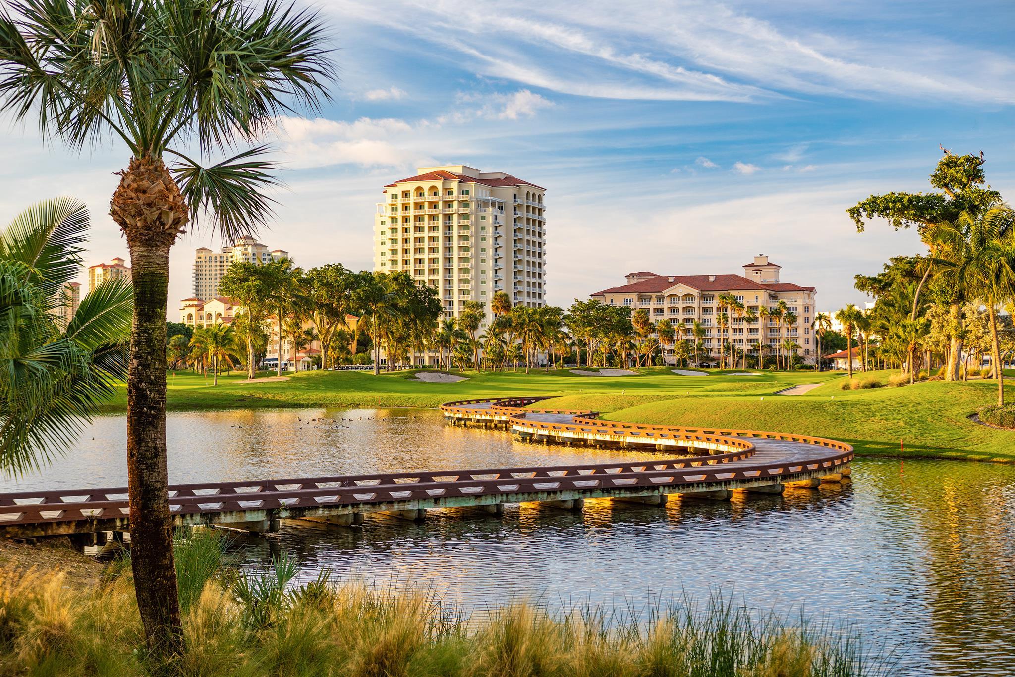 A beautiful golf course with a wooden bridge curving over the water, surrounded by palm trees and tall buildings in the background.