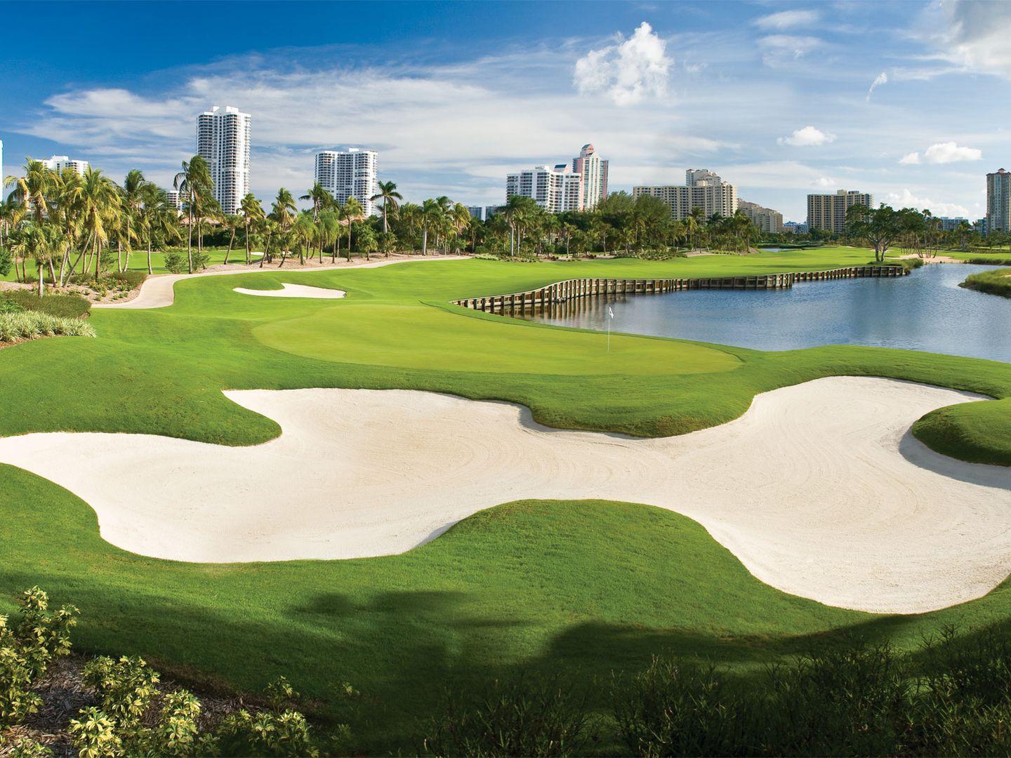 A pristine golf hole with large white sand bunkers and a water hazard, framed by tropical palm trees and city buildings.
