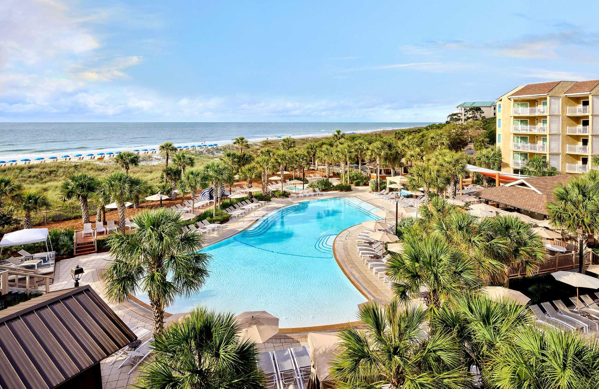 Aerial view of the swimming pool surrounded by palm trees with coastal views in the distance