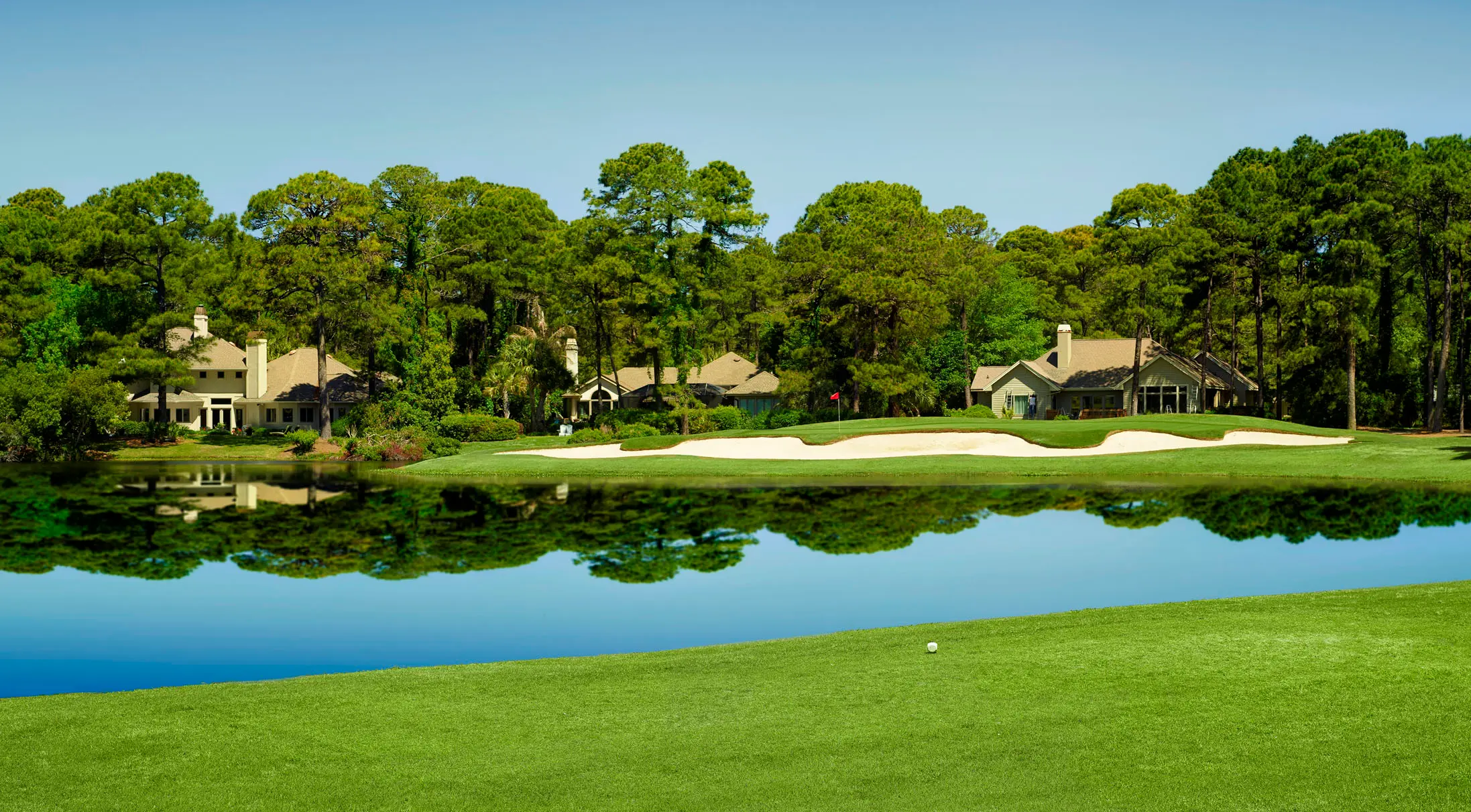 A serene golf course with a large sand trap and calm reflective water.