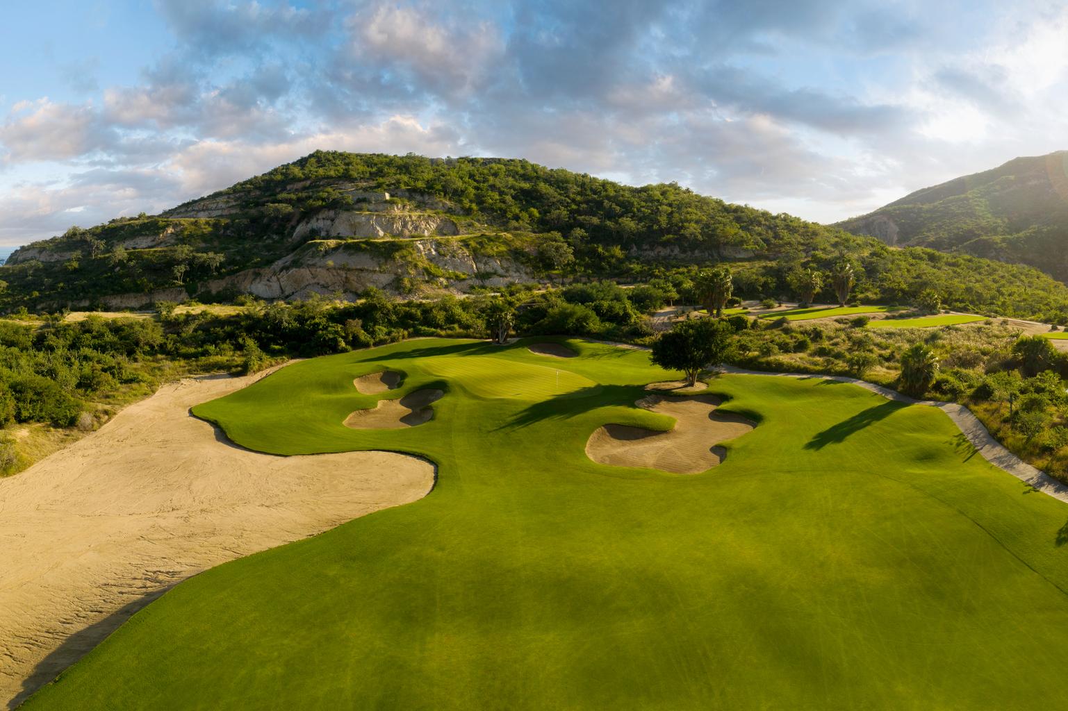A scenic golf course surrounded by rugged hills and lush greenery, featuring sand bunkers and smooth greens.