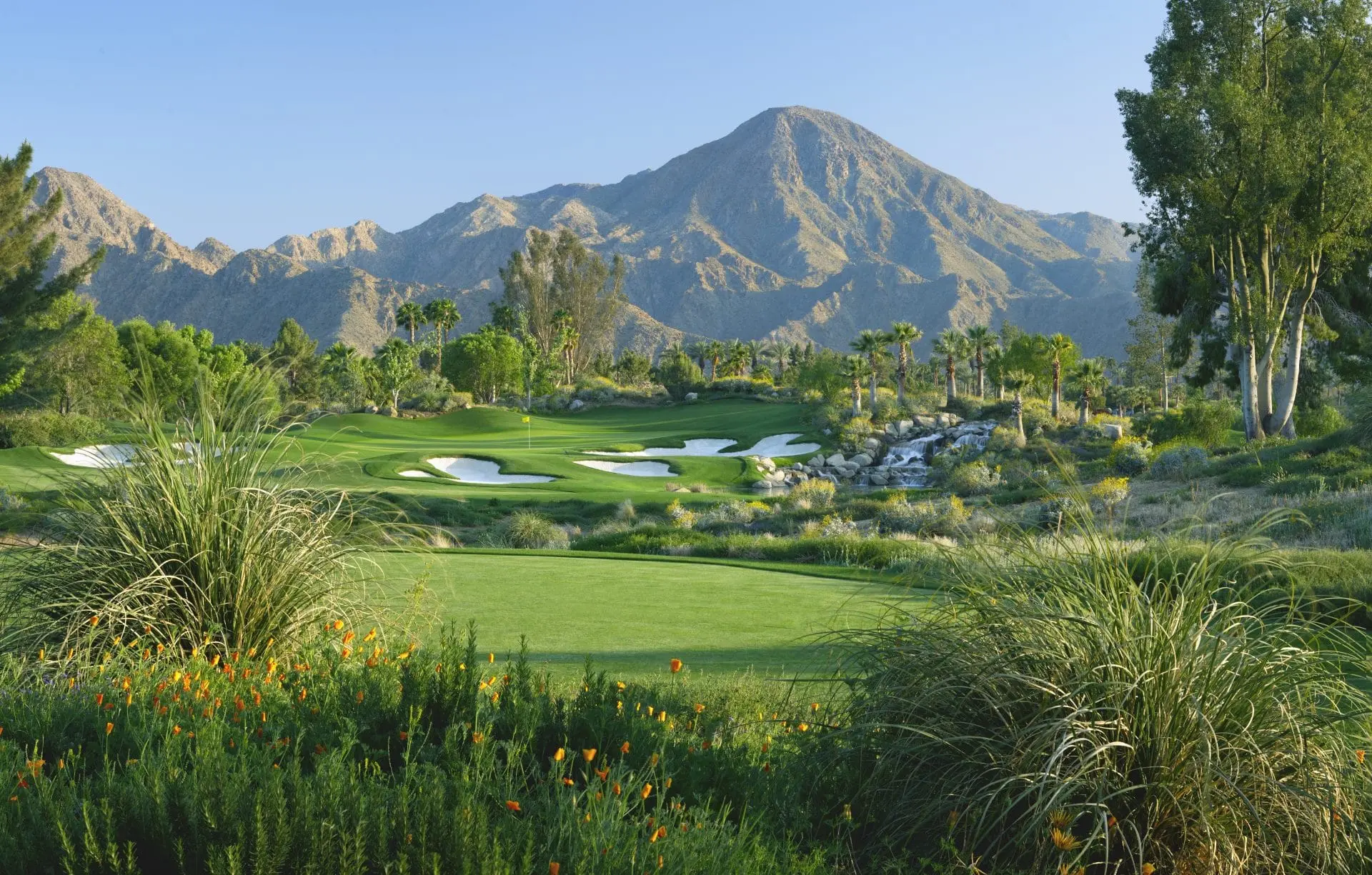 A pristine course with lush greenery, white sand bunkers, and a backdrop of rocky desert peaks.