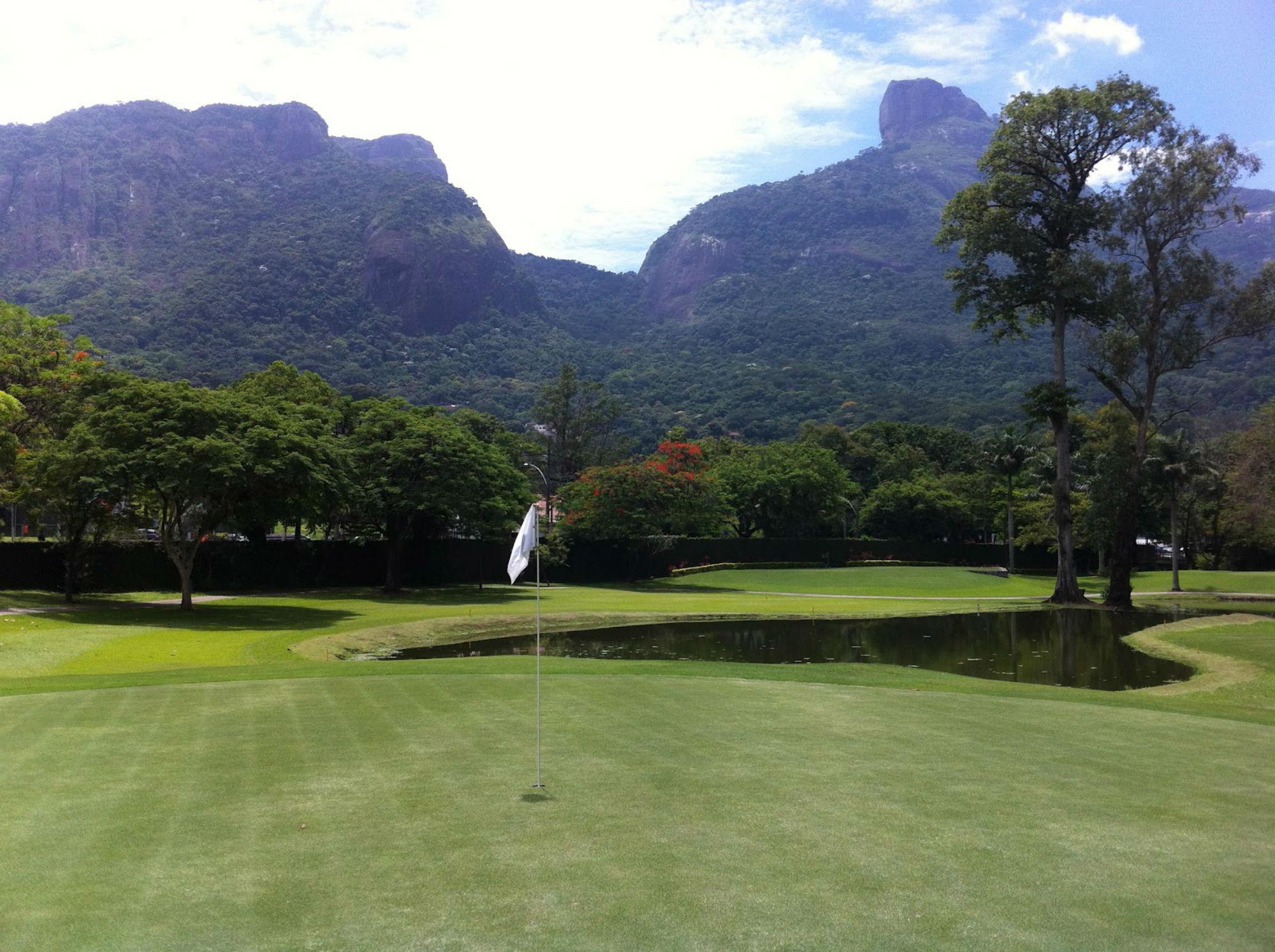 A green golf course with a flag near a small pond, framed by mountains and dense trees