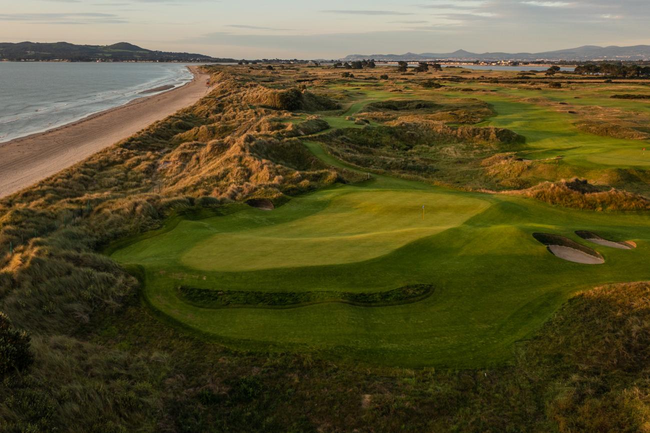 A coastal golf course with a green near the beach, surrounded by sandy dunes.